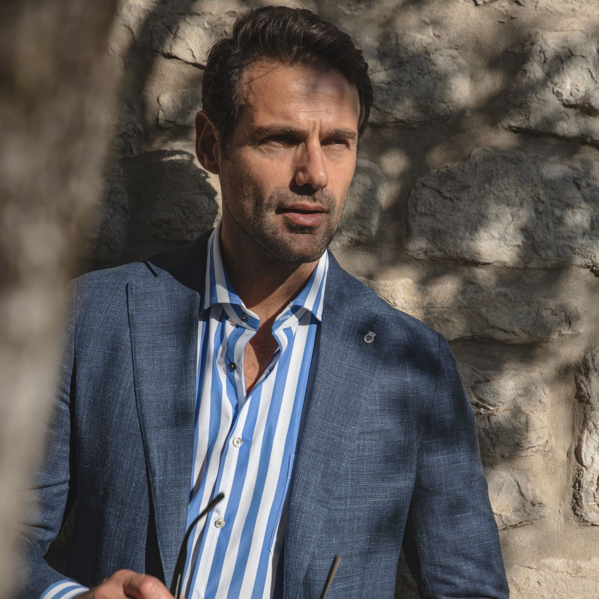 Man in blue blazer and striped shirt leans against stone wall, holding glasses.