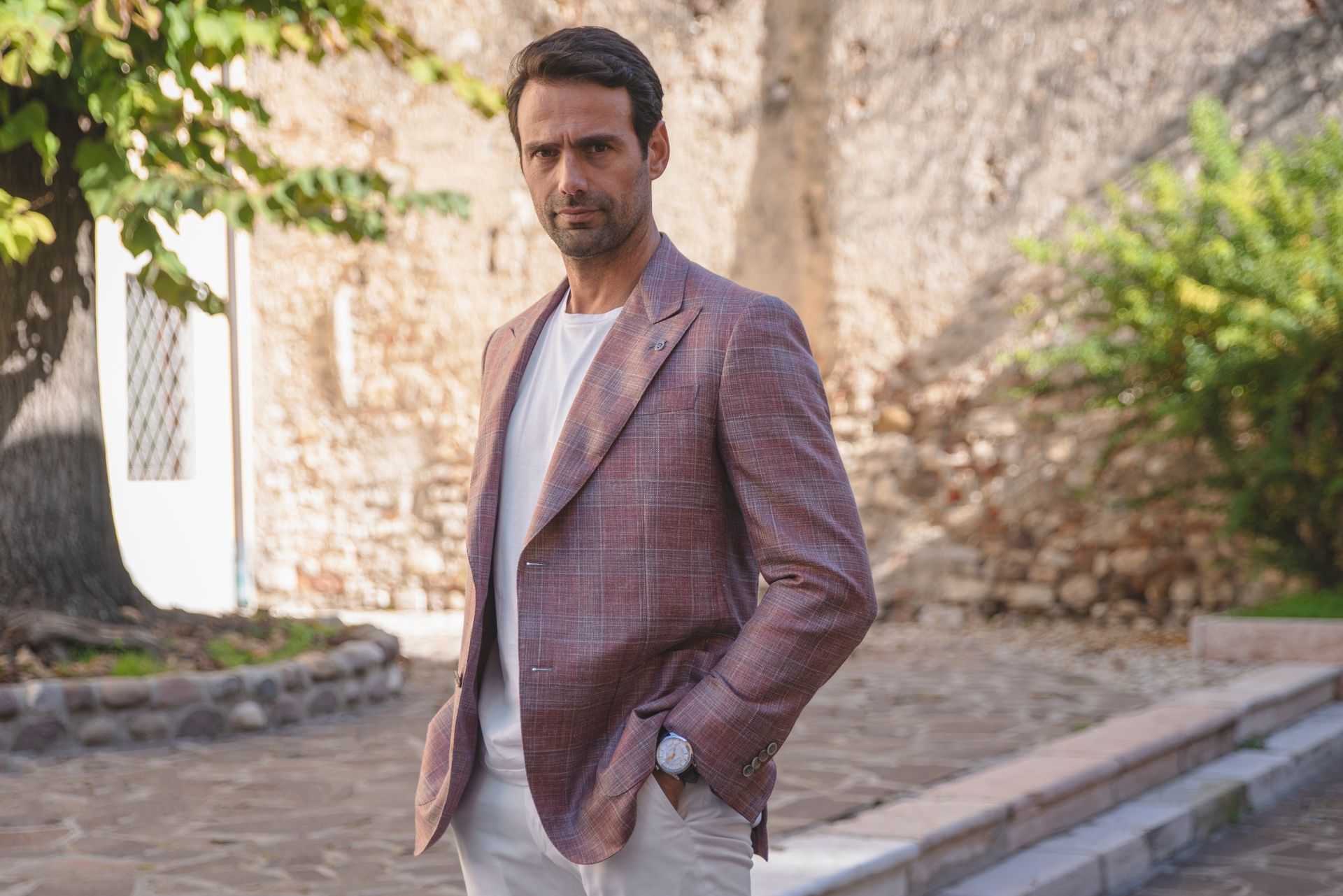 Man in plaid blazer and white shirt stands outdoors with hands in pockets.