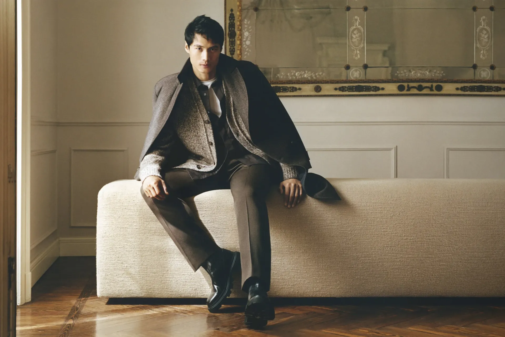 Man seated on a bench, wearing a coat, brown pants, and boots. Interior setting with a mirror.