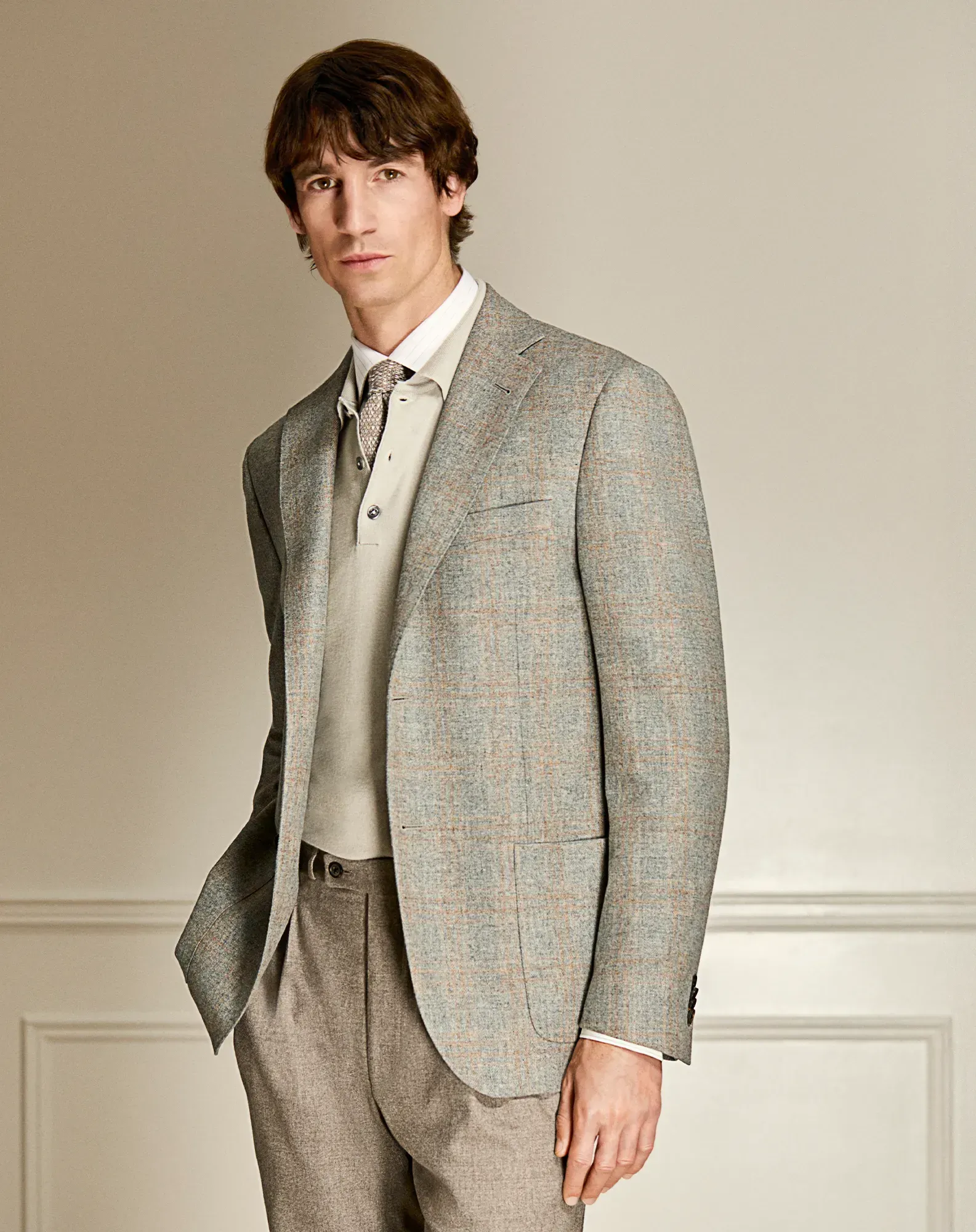 Man in tweed blazer, light sweater, and pleated trousers, leaning against a wall.