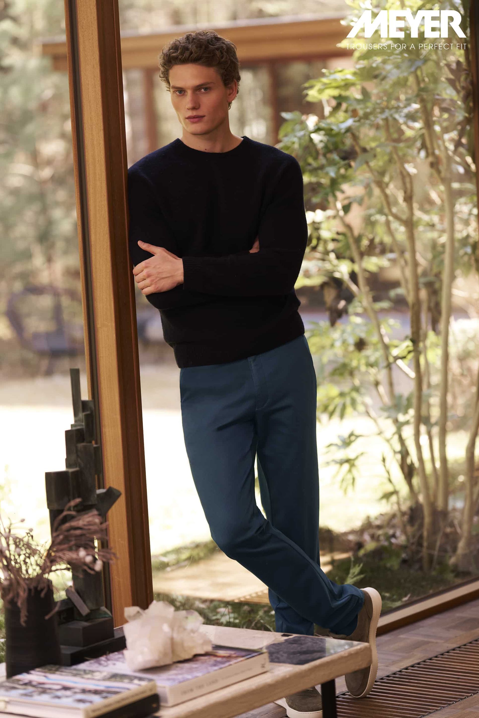 Man in blue pants and black sweater leans against a window. Outdoor setting with greenery.