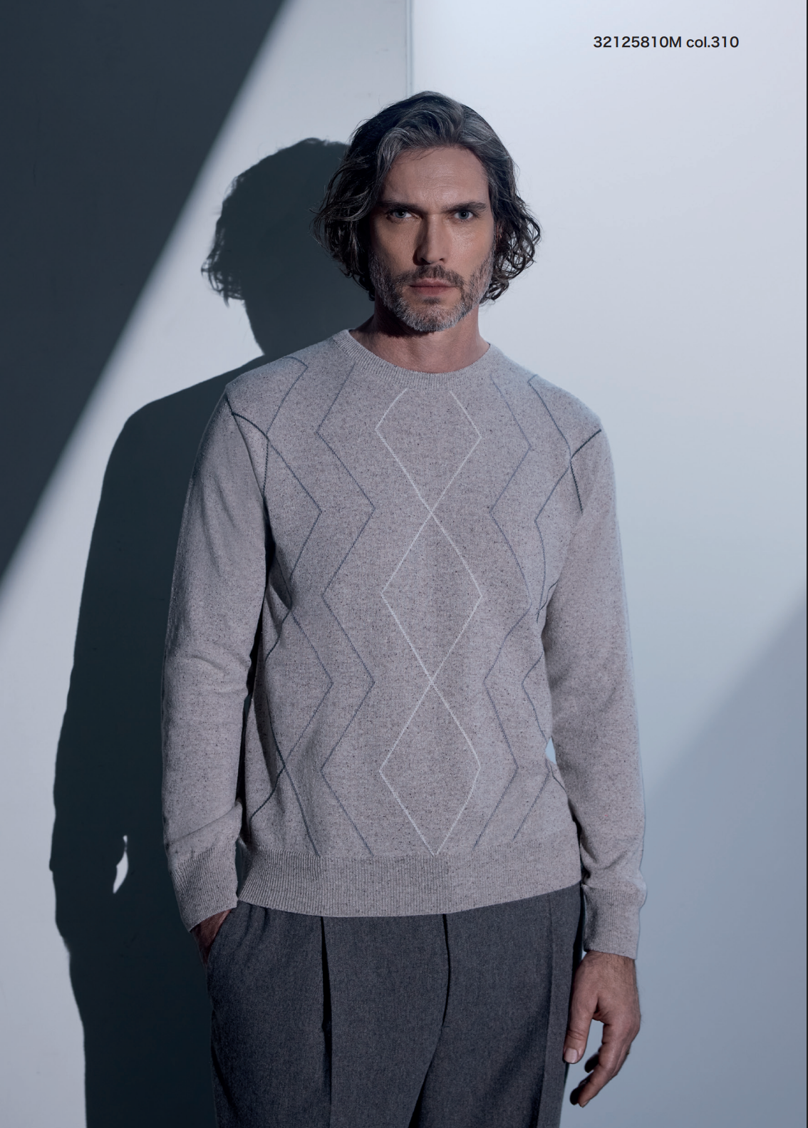 Man in gray sweater and trousers, standing against a white backdrop, hands in pockets.