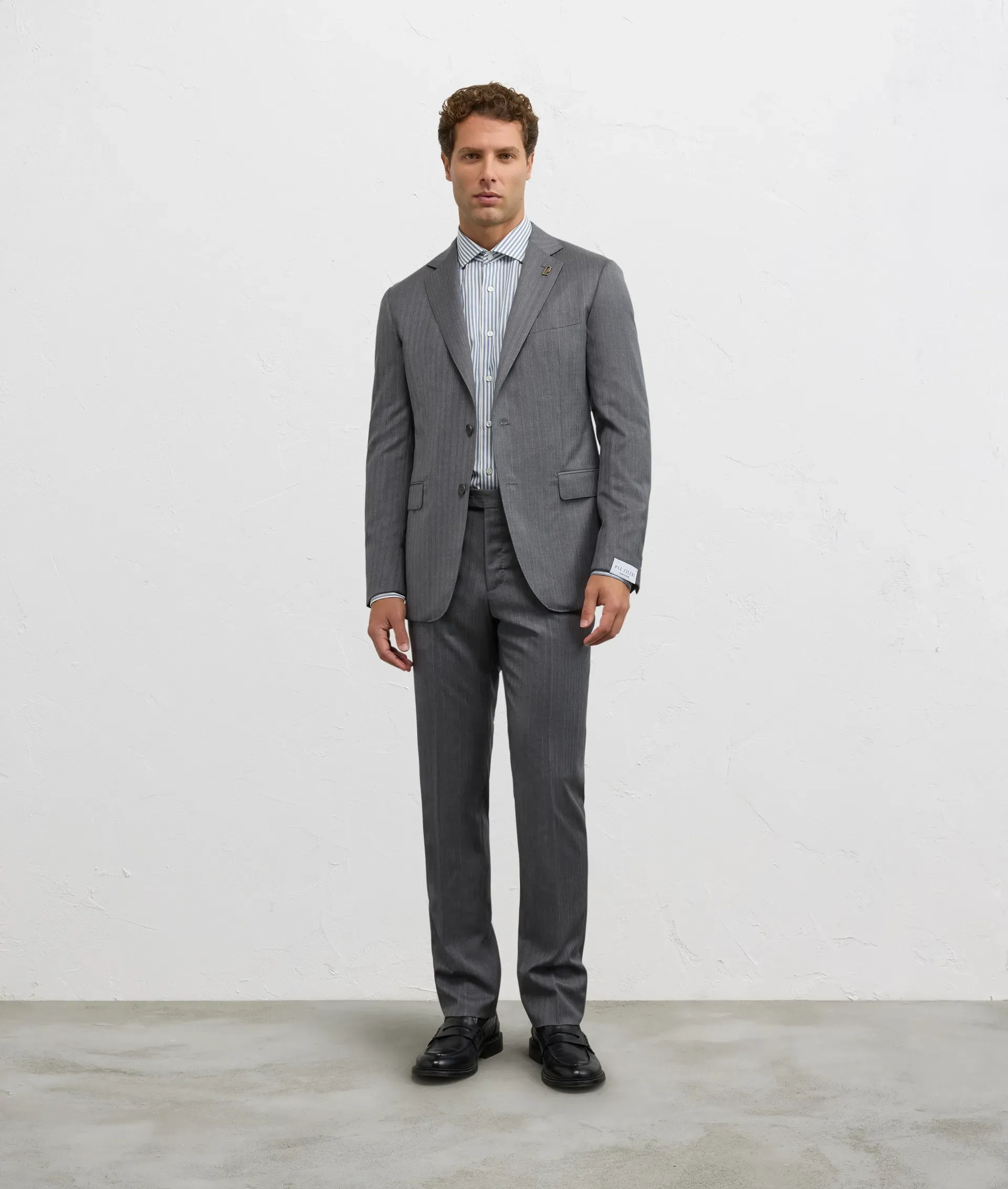 Man in a gray suit and striped shirt standing against a white wall.