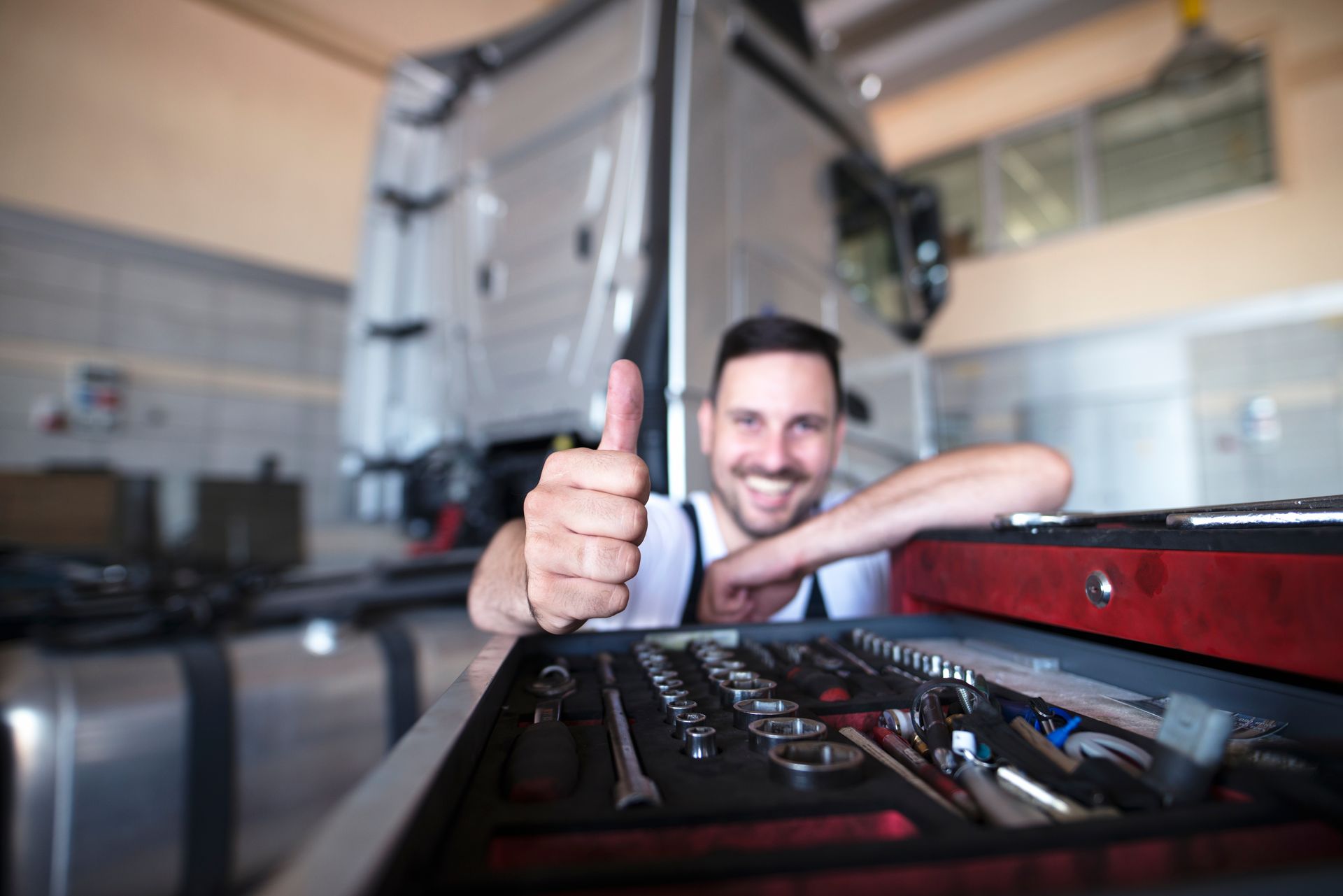 Mechanic gives a thumbs up, smiling, in front of a truck with a toolbox in view, in a garage setting.