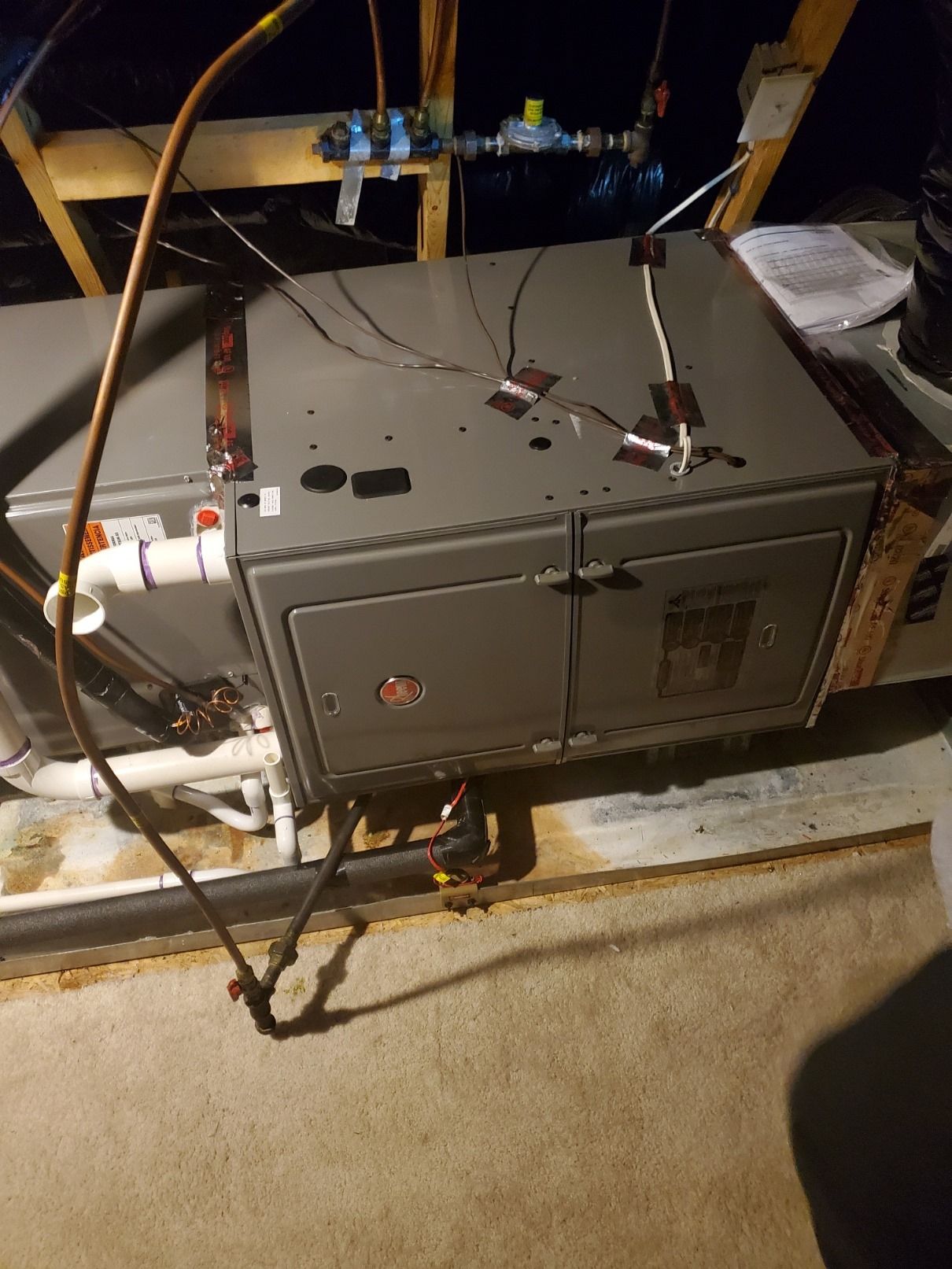 A Rheem HVAC unit in an attic with copper tubing and wiring visible, sitting on a concrete floor.