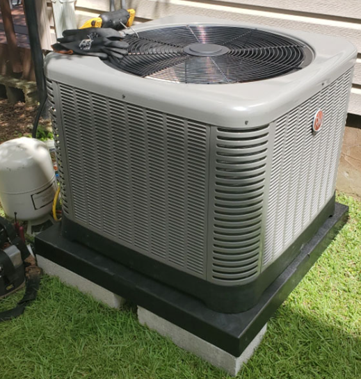 Gray air conditioning unit on a black base, sitting on concrete blocks on grass.