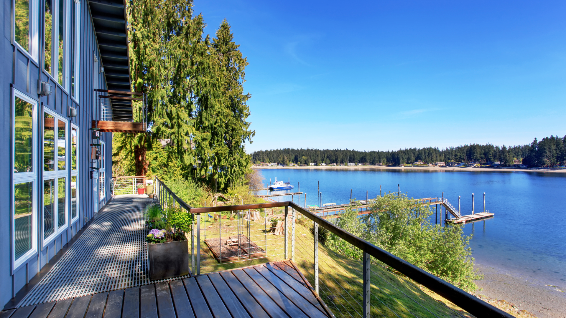 Sunlit deck beside a blue waterfront, with railings, trees, and a distant shoreline under a clear sky