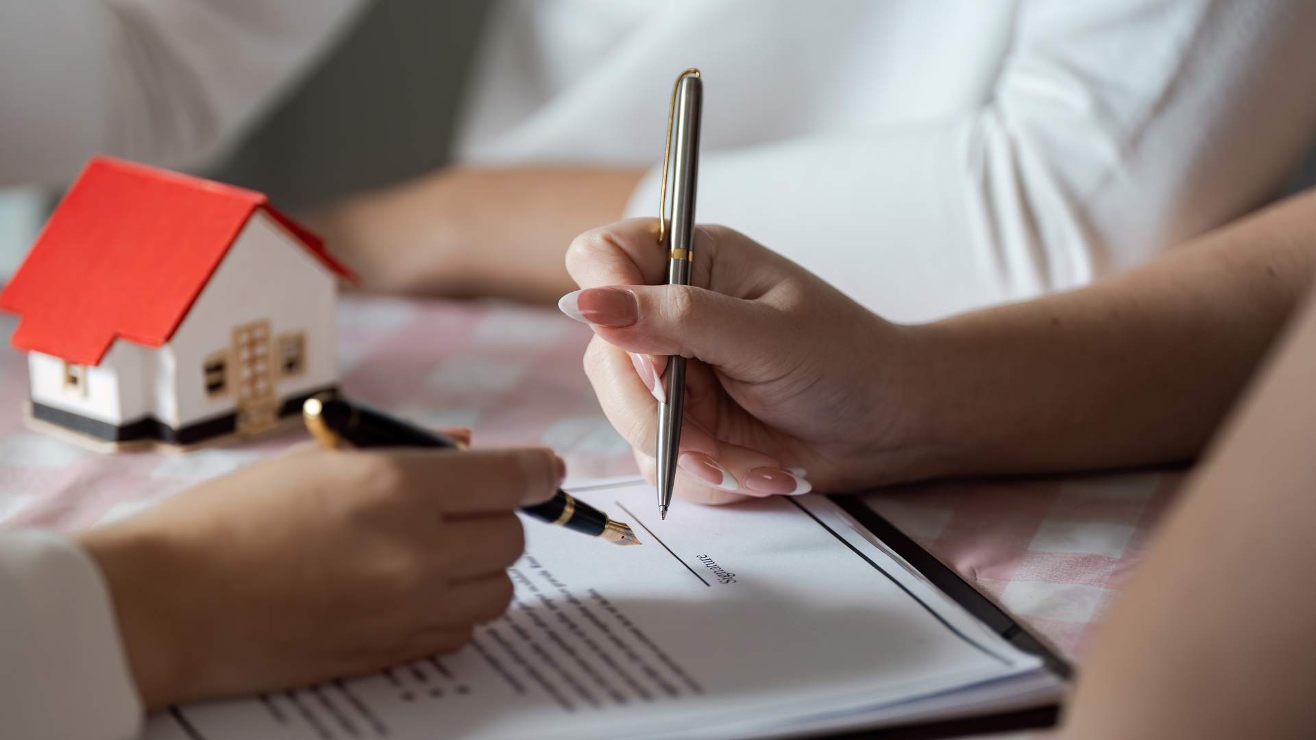 Hands signing a document beside a small model house on a desk