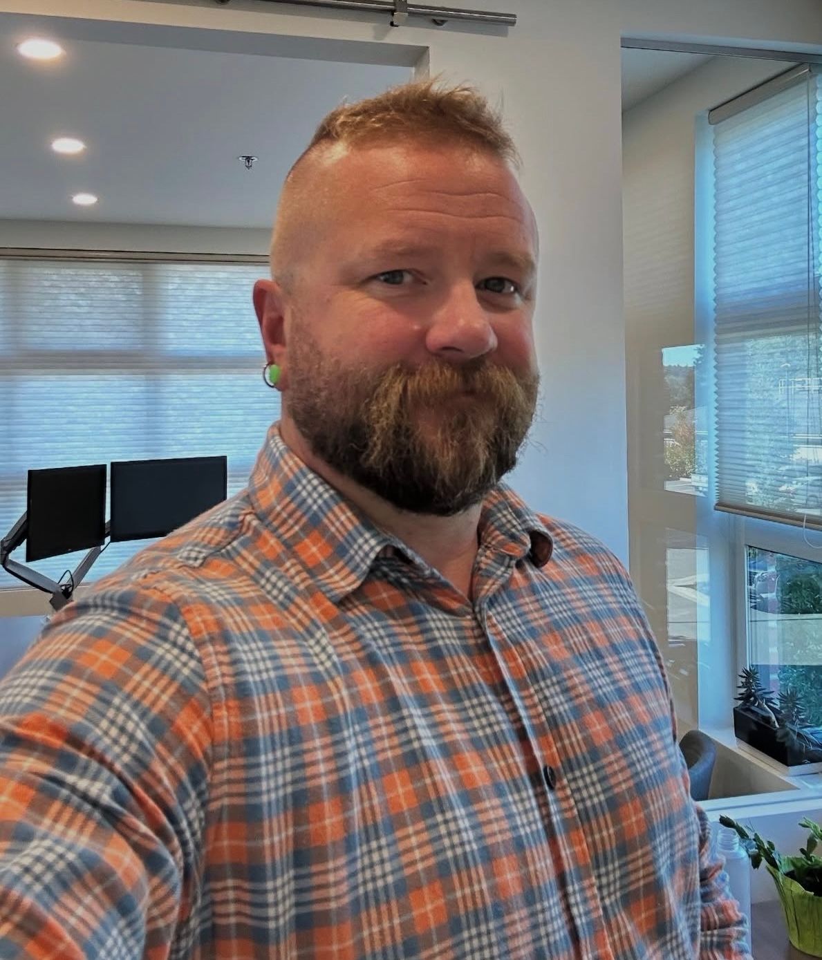 Smiling man in plaid shirt in a bright office with windows and blinds.