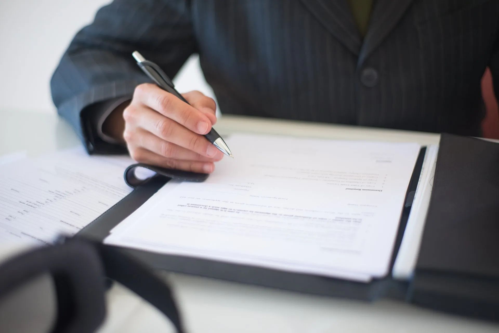A person in a suit writing on a document in a binder with a pen at a desk.