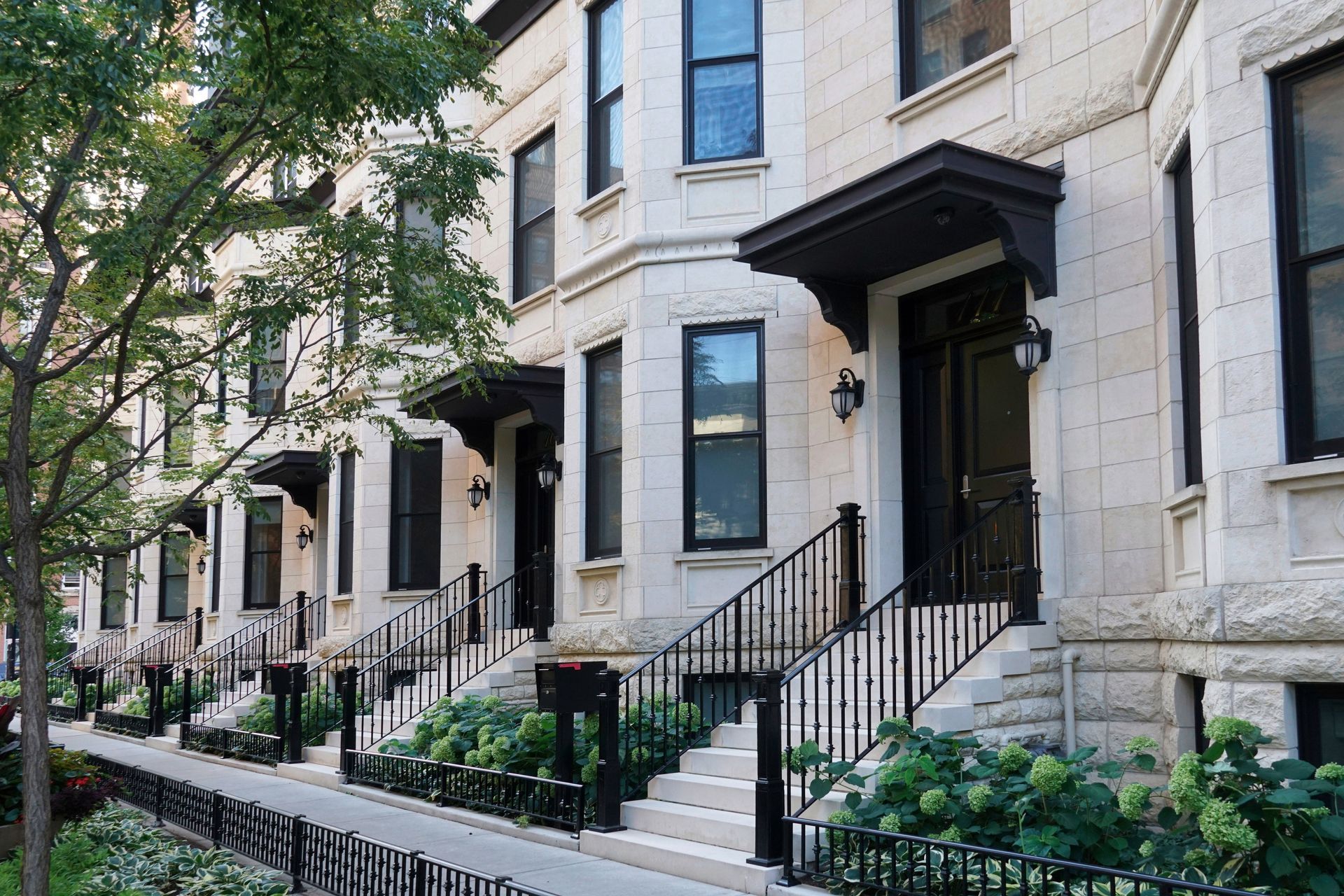 Row of light stone townhouses with black doors and railings; lush green bushes line the stairs.