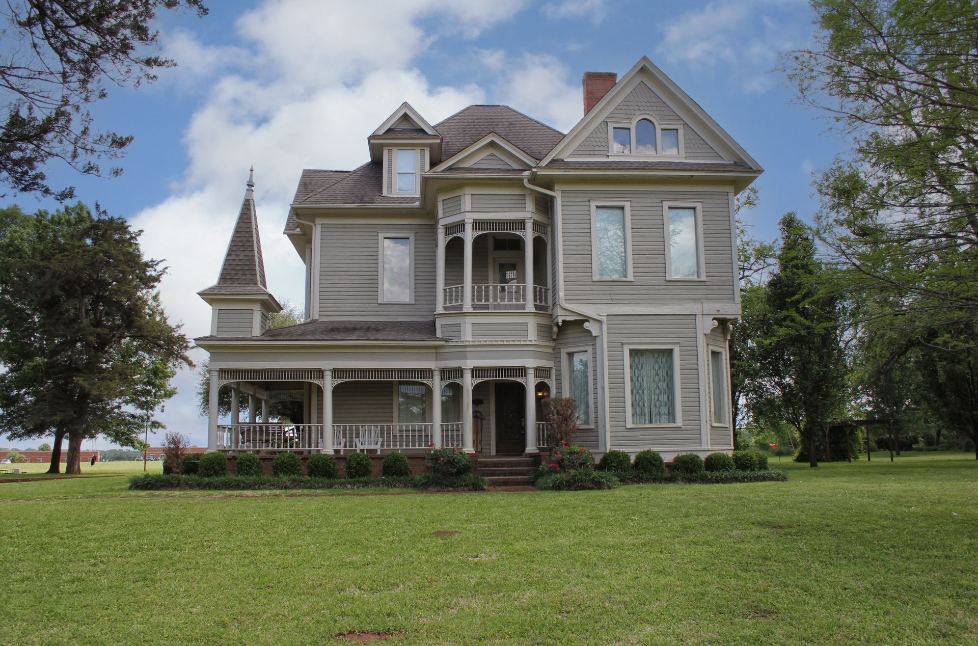 Victorian-style gray house with a wraparound porch, set on a grassy lawn under a partly cloudy sky.