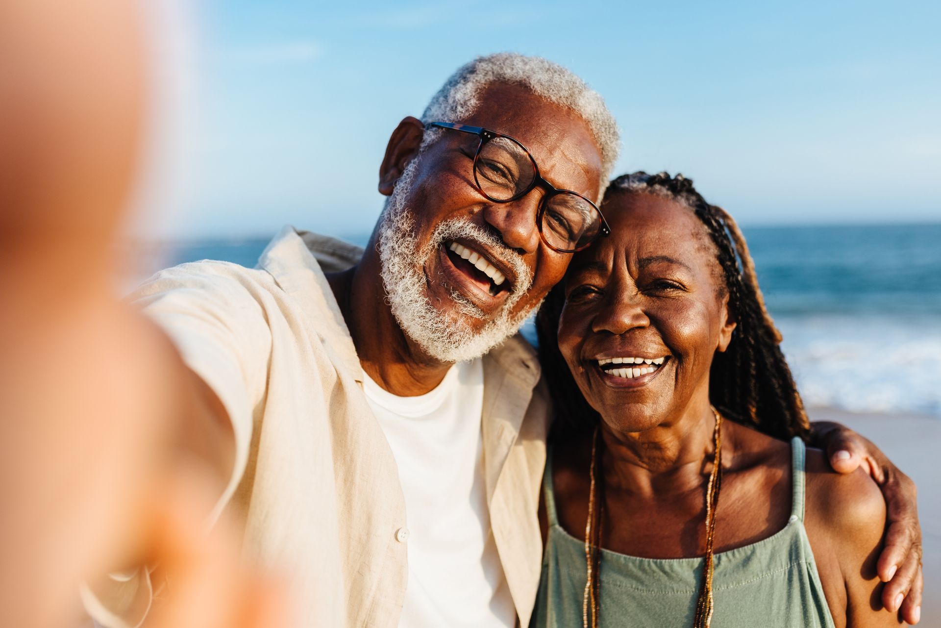 An elderly Black couple smiles widely while taking a selfie on a beach, ocean in the background.