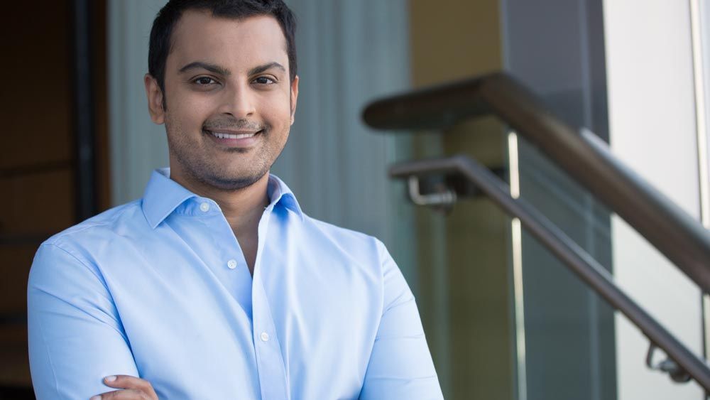 A man in a blue shirt is standing with his arms crossed and smiling.