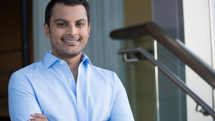 A man in a blue shirt is standing with his arms crossed and smiling.