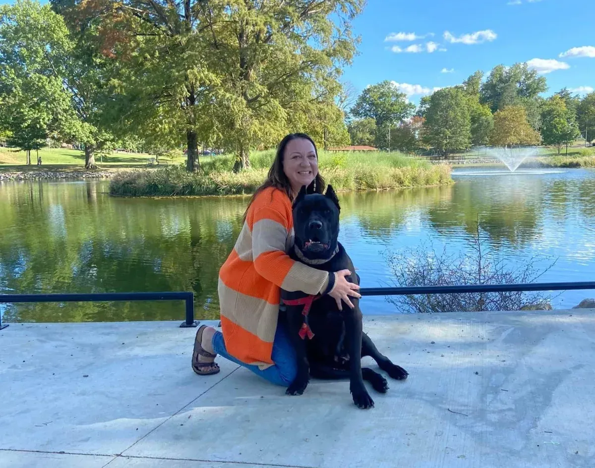 A woman is kneeling down next to a black dog in front of a lake.