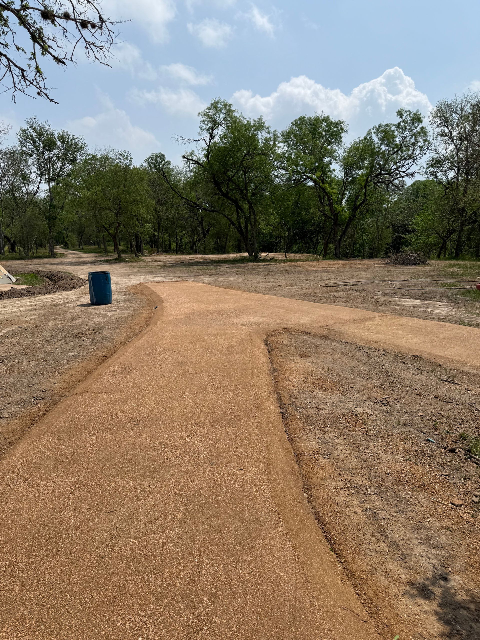 A gravel path splitting into two directions in a park, with trees and a blue barrel visible.