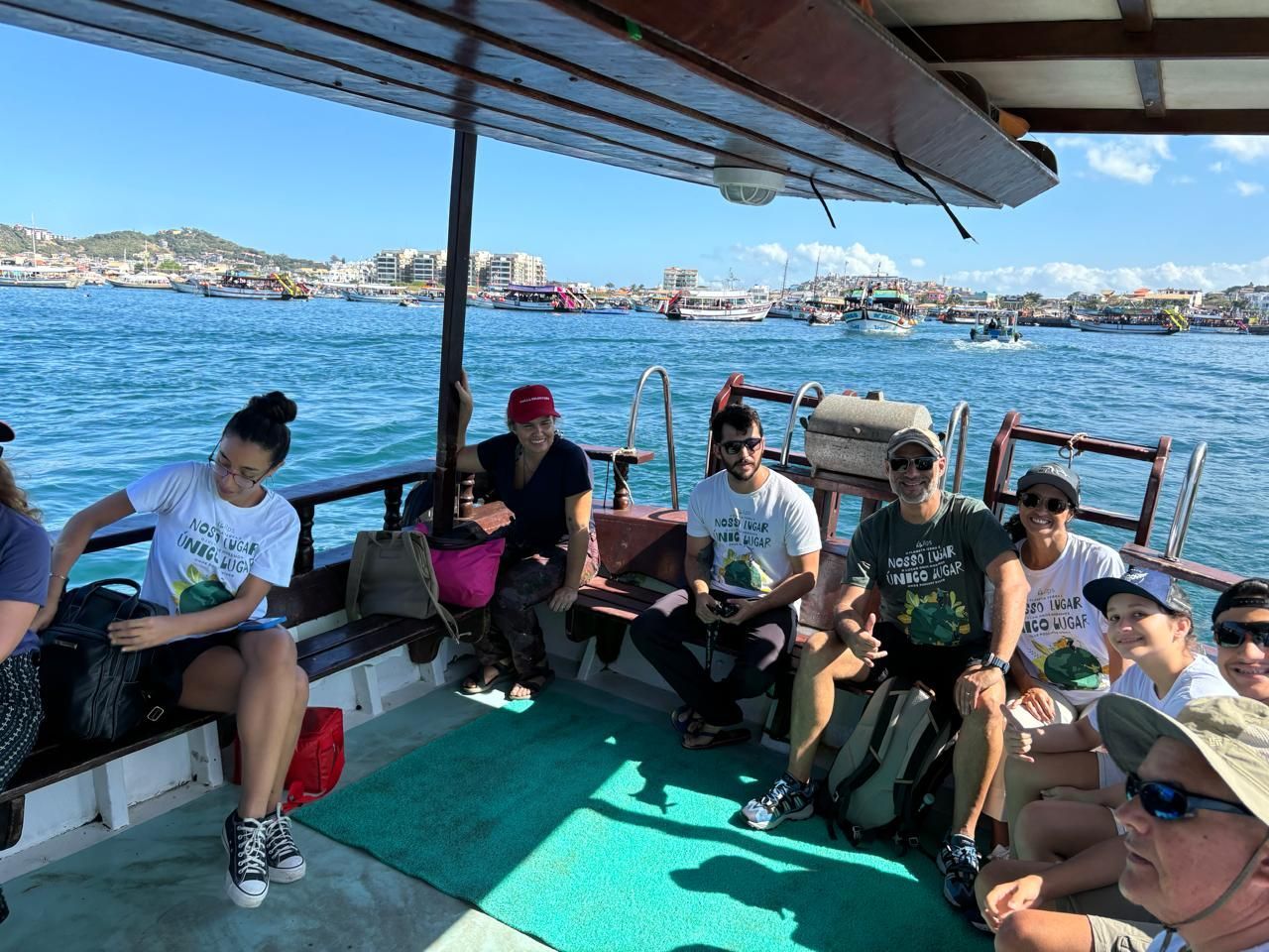 Pessoas em um barco aproveitando um dia ensolarado no mar; água azul-turquesa e uma paisagem urbana ao fundo.