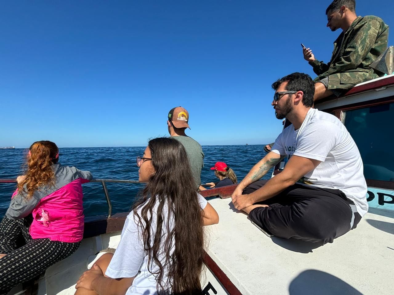 Pessoas em um barco em um dia ensolarado, olhando para o oceano. Algumas estão sentadas, outras em pé, observando.