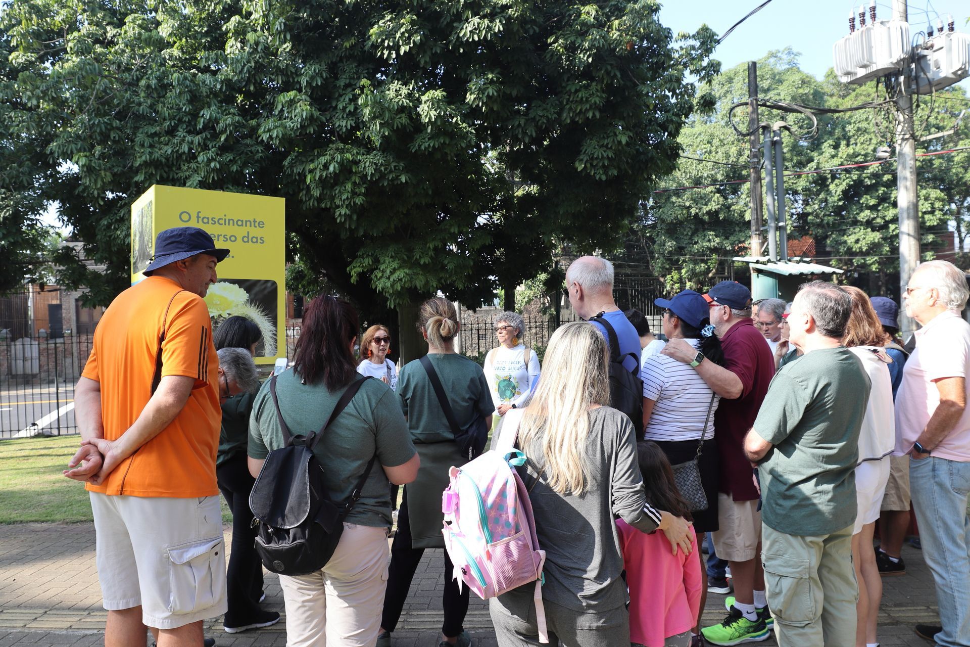 Um grupo de pessoas ouve alguém perto de uma placa amarela sob as árvores em um ambiente externo.