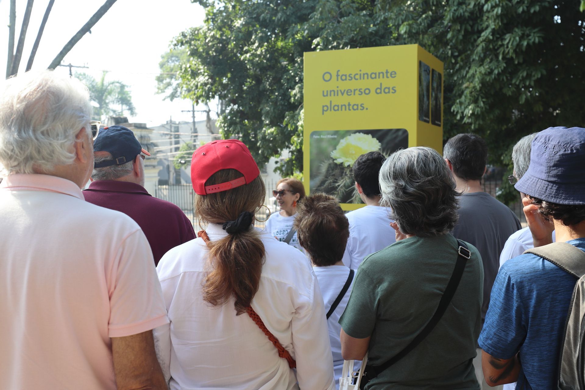 Um grupo de pessoas reunidas ao ar livre, ouvindo uma mulher em frente a um painel amarelo. O painel tem um texto e uma foto de uma flor.