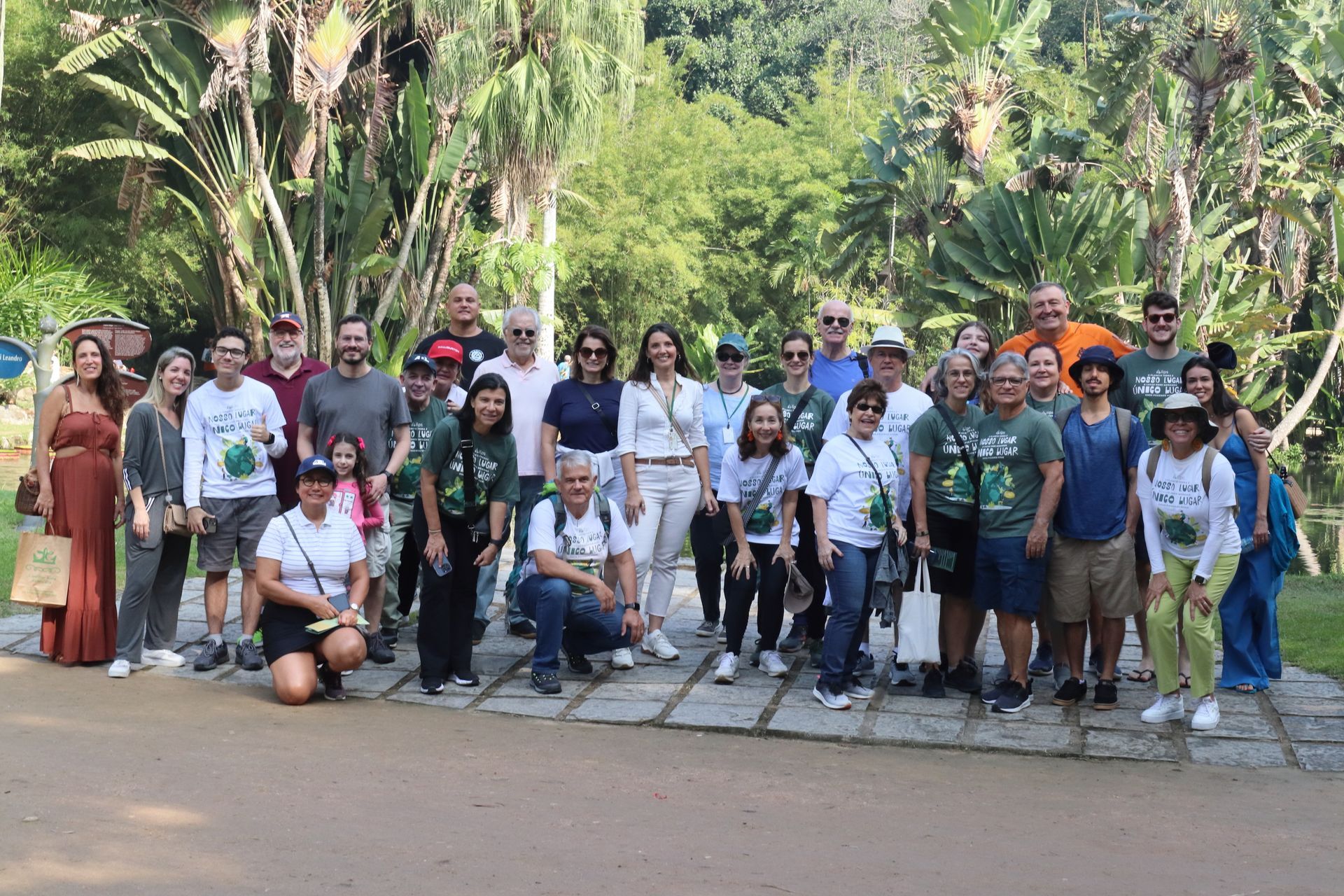 Grupo de pessoas sorrindo para uma foto ao ar livre, em um parque verdejante. Alguns usam camisetas brancas com um logotipo, outros estão vestidos casualmente.