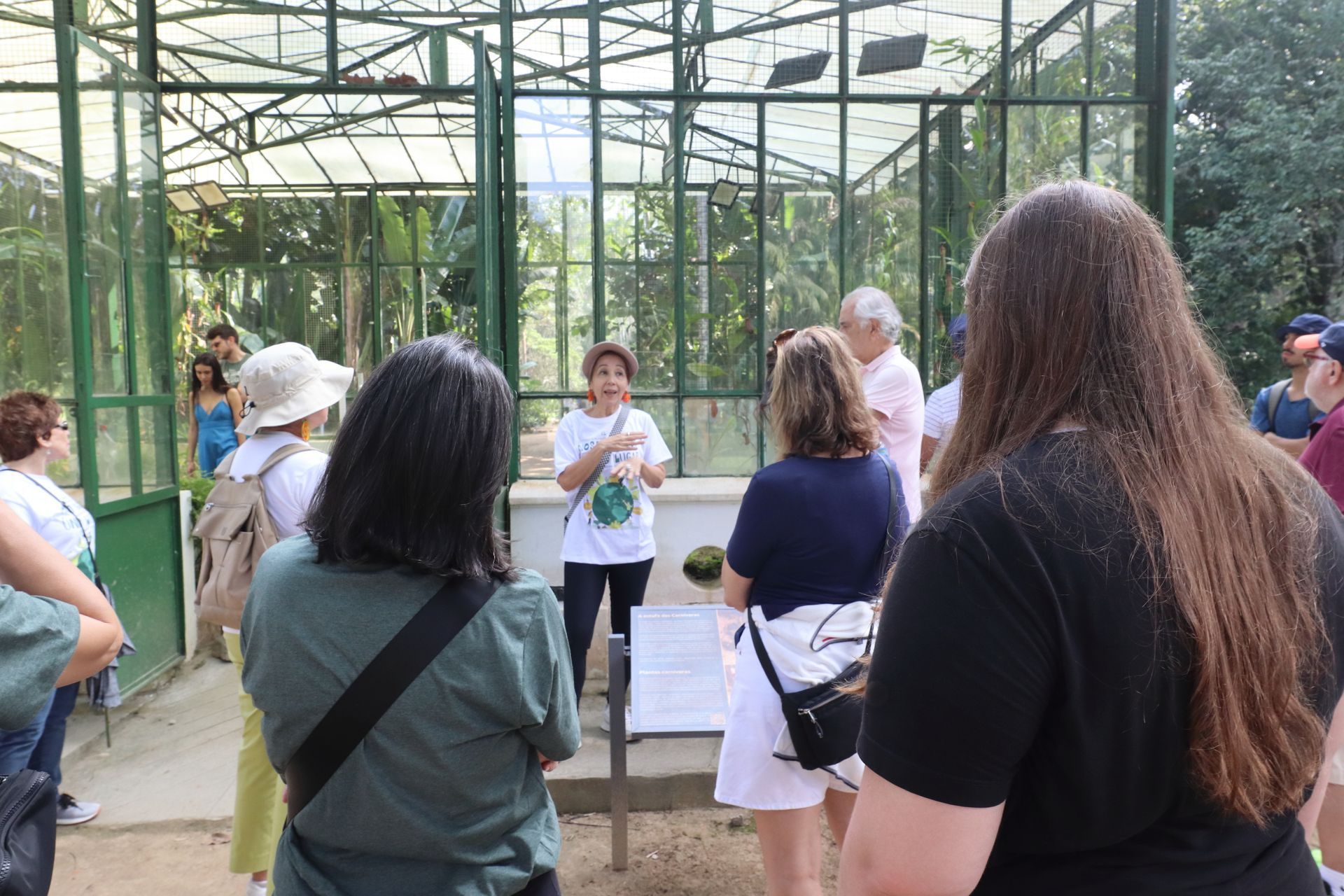 Um grupo de pessoas ouve uma mulher fazendo uma apresentação dentro de uma estufa com paredes de vidro. O grupo está ao ar livre, sob a luz do dia.