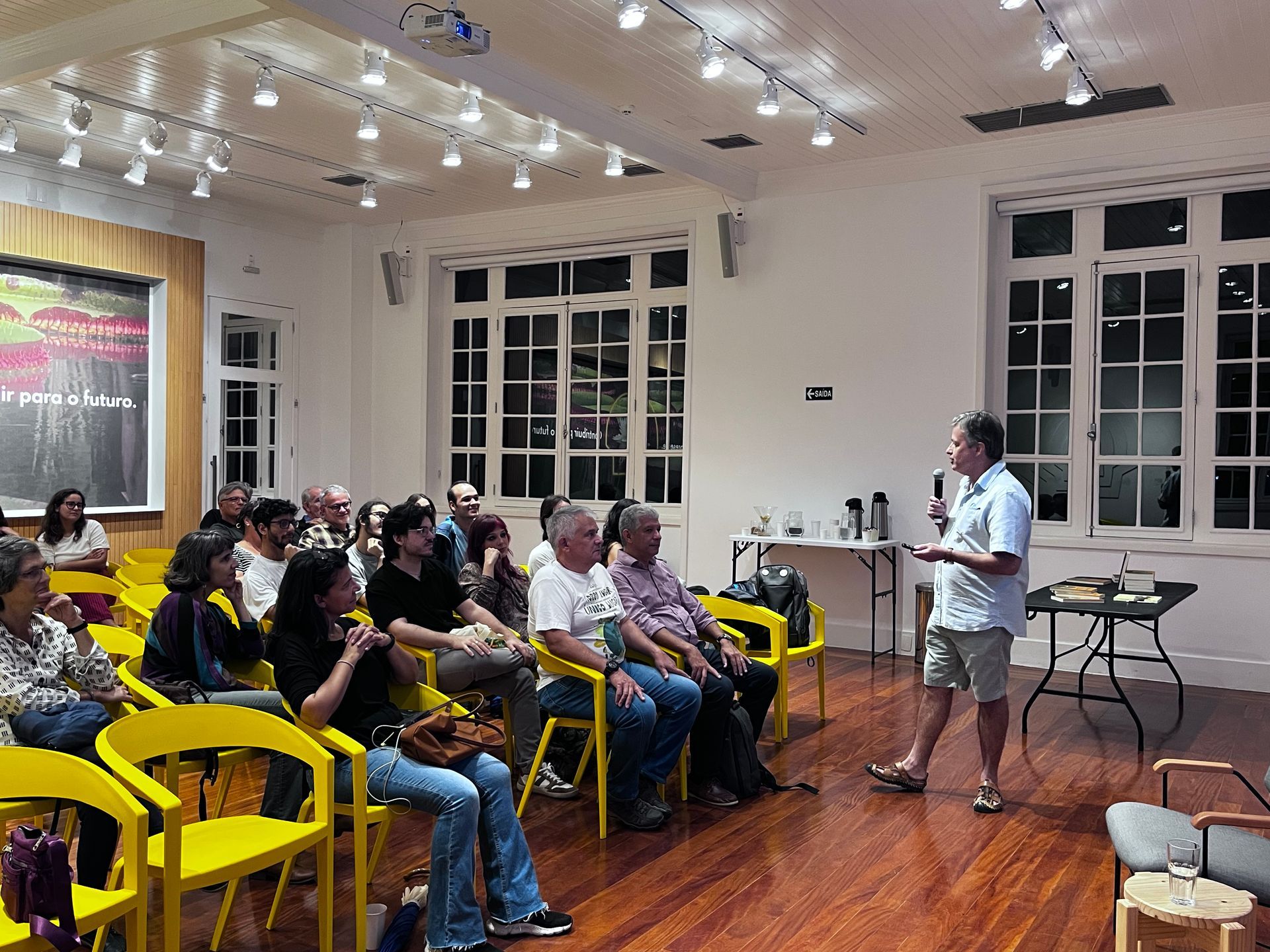 Um homem fala ao microfone para uma plateia sentada em cadeiras amarelas. A sala tem janelas grandes e piso de madeira.