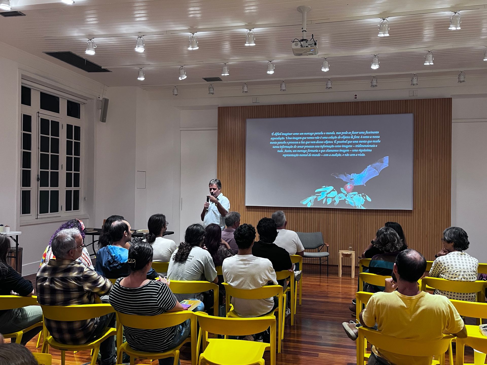 Um homem apresenta para uma plateia sentada em uma sala bem iluminada com uma tela exibindo um gráfico. Cadeiras amarelas estão de frente para o apresentador.
