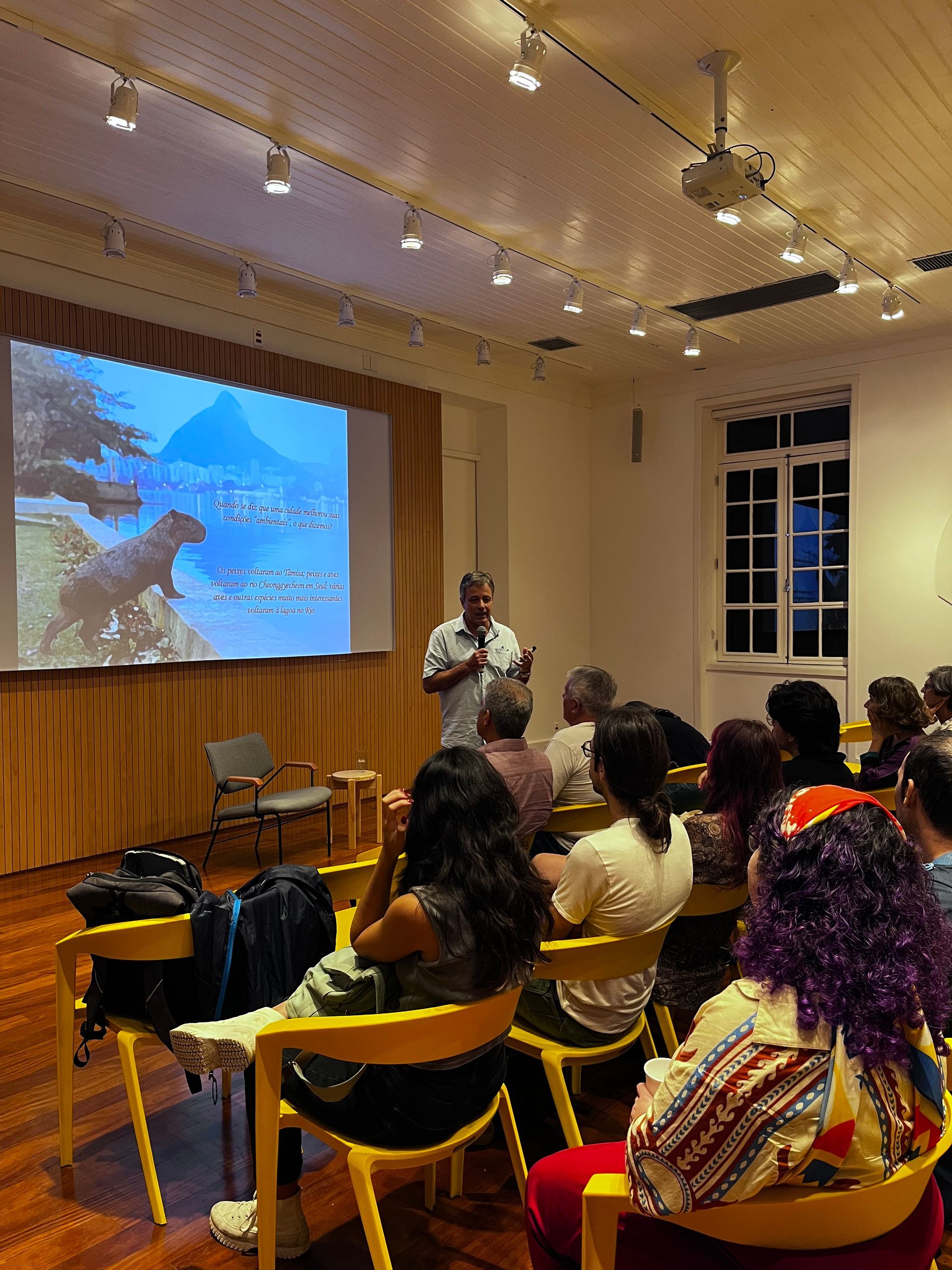 Um homem faz uma apresentação para uma plateia em uma sala iluminada. Uma tela de projetor mostra uma paisagem. O público está sentado em cadeiras amarelas.