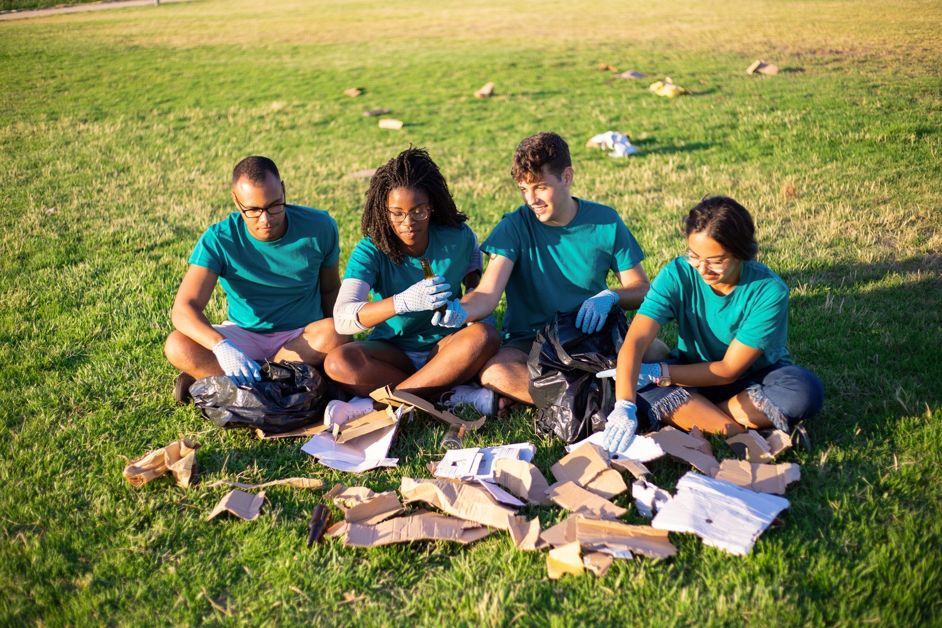 Four people in teal shirts sit on grass, sorting papers outdoors in sunlight.