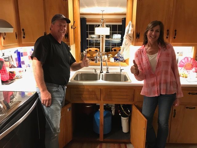 A man and a woman are standing in front of a kitchen sink giving a thumbs up