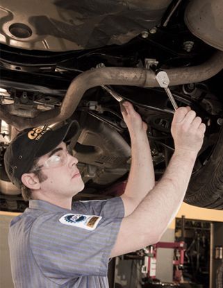 Mechanic working under a car, using a wrench. Wearing glasses and a cap, in a garage setting.