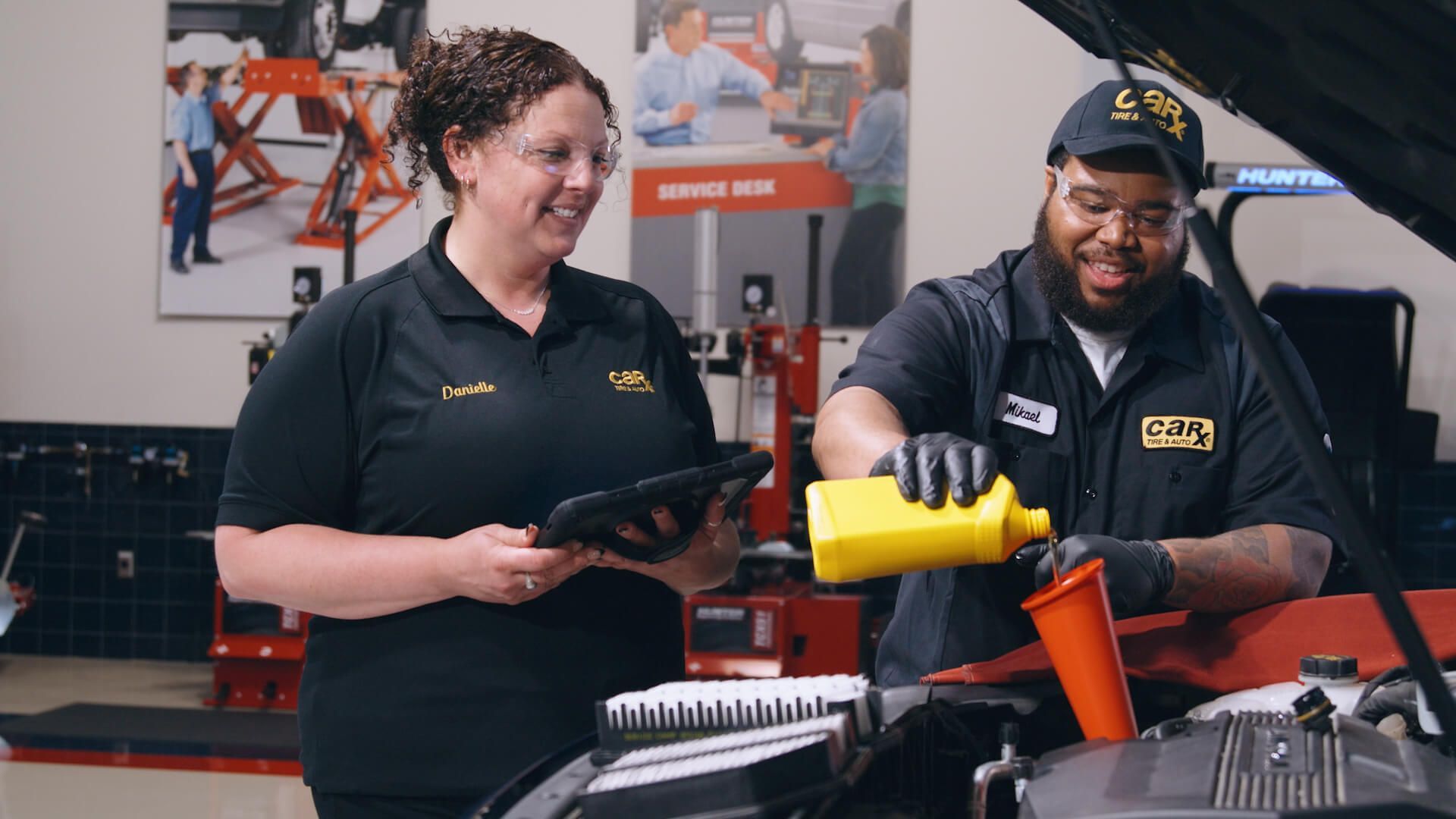 Two auto technicians working on a car, pouring oil. One holds a tablet, smiling. In a repair shop.
