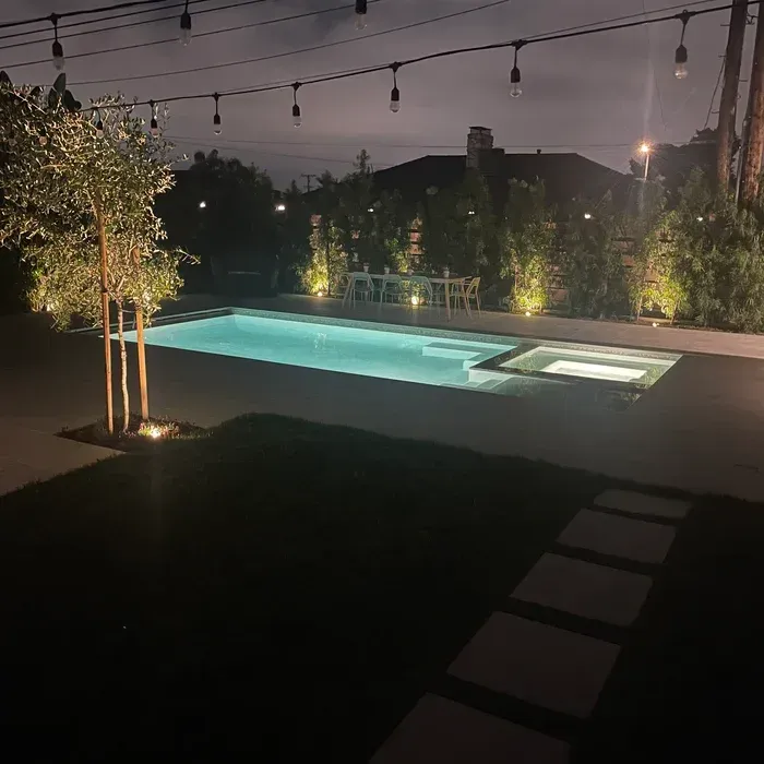 Nighttime backyard pool with glowing turquoise water, string lights overhead, and stepping stones through the lawn