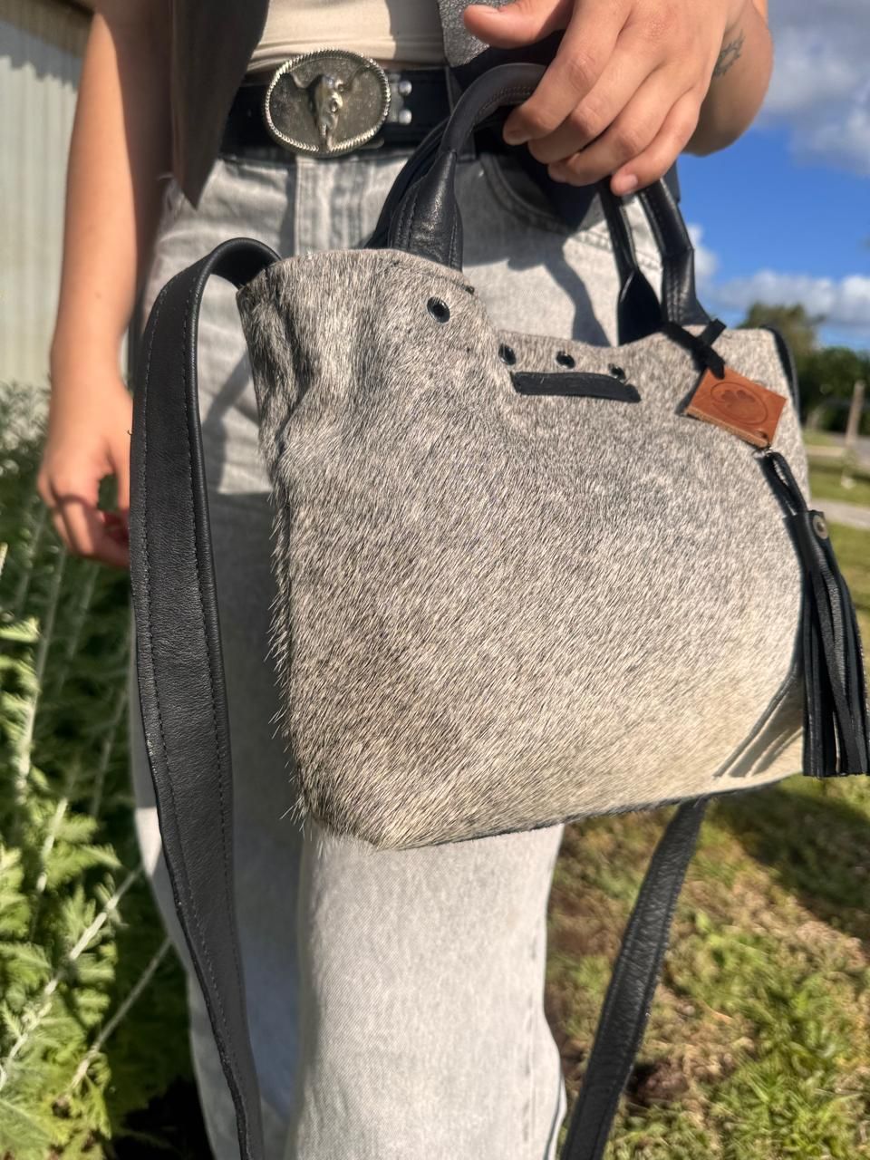 Mano sosteniendo un pequeño bolso de cuero beige texturizado con asas y correa negras, sobre un fondo verde desenfocado.