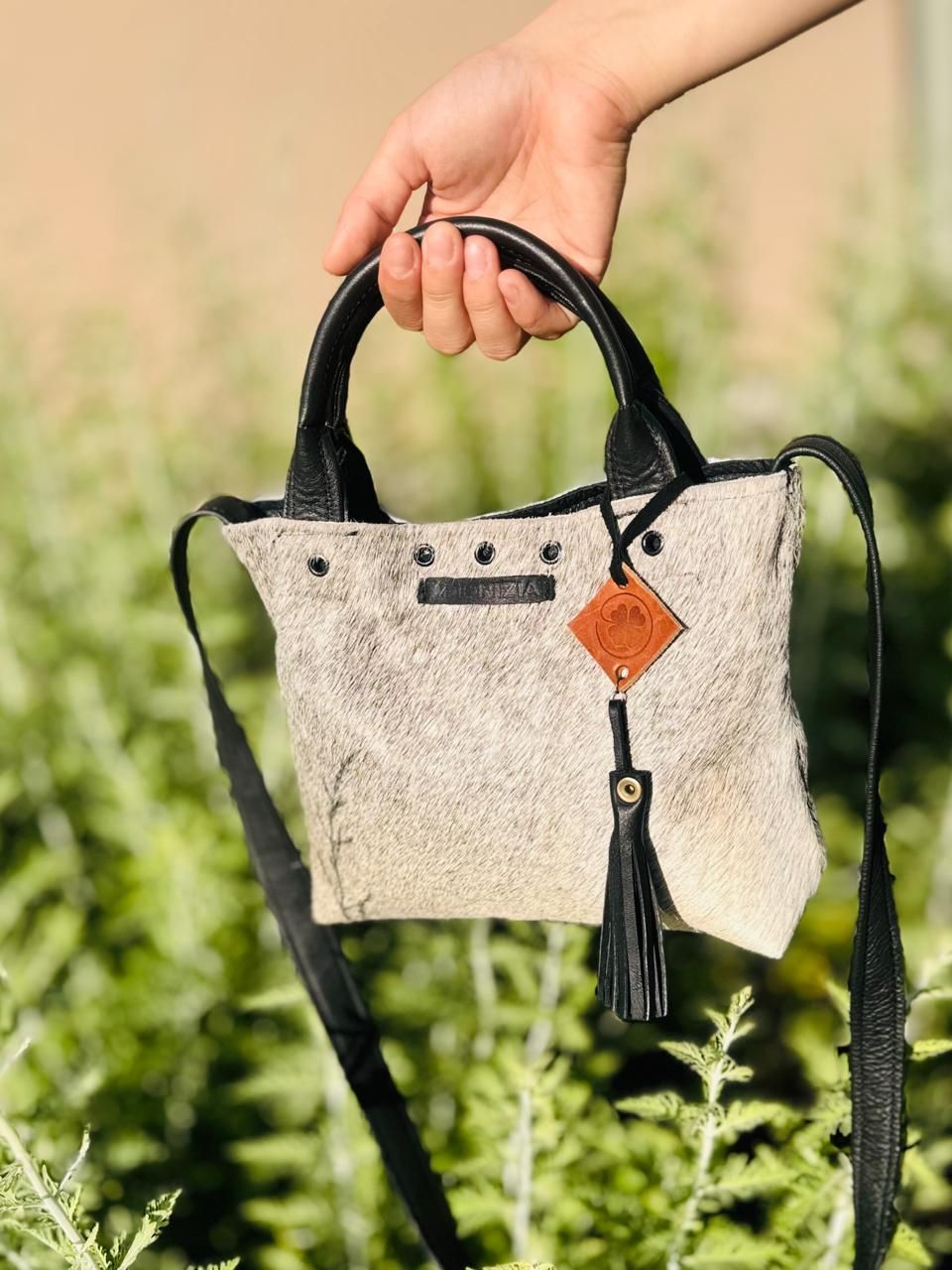 Mano sosteniendo un pequeño bolso de mano de piel texturizada en tonos beige y negro, con correa cruzada y borla.