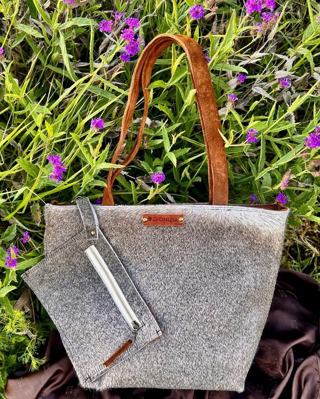 Bolso tote gris con asas de cuero marrón y pequeño neceser a juego en un entorno de jardín.