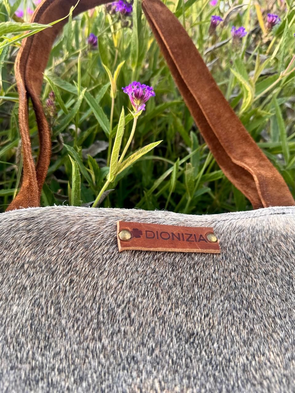 Bolso tote gris con asas de cuero marrón y pequeño neceser a juego en un entorno de jardín.