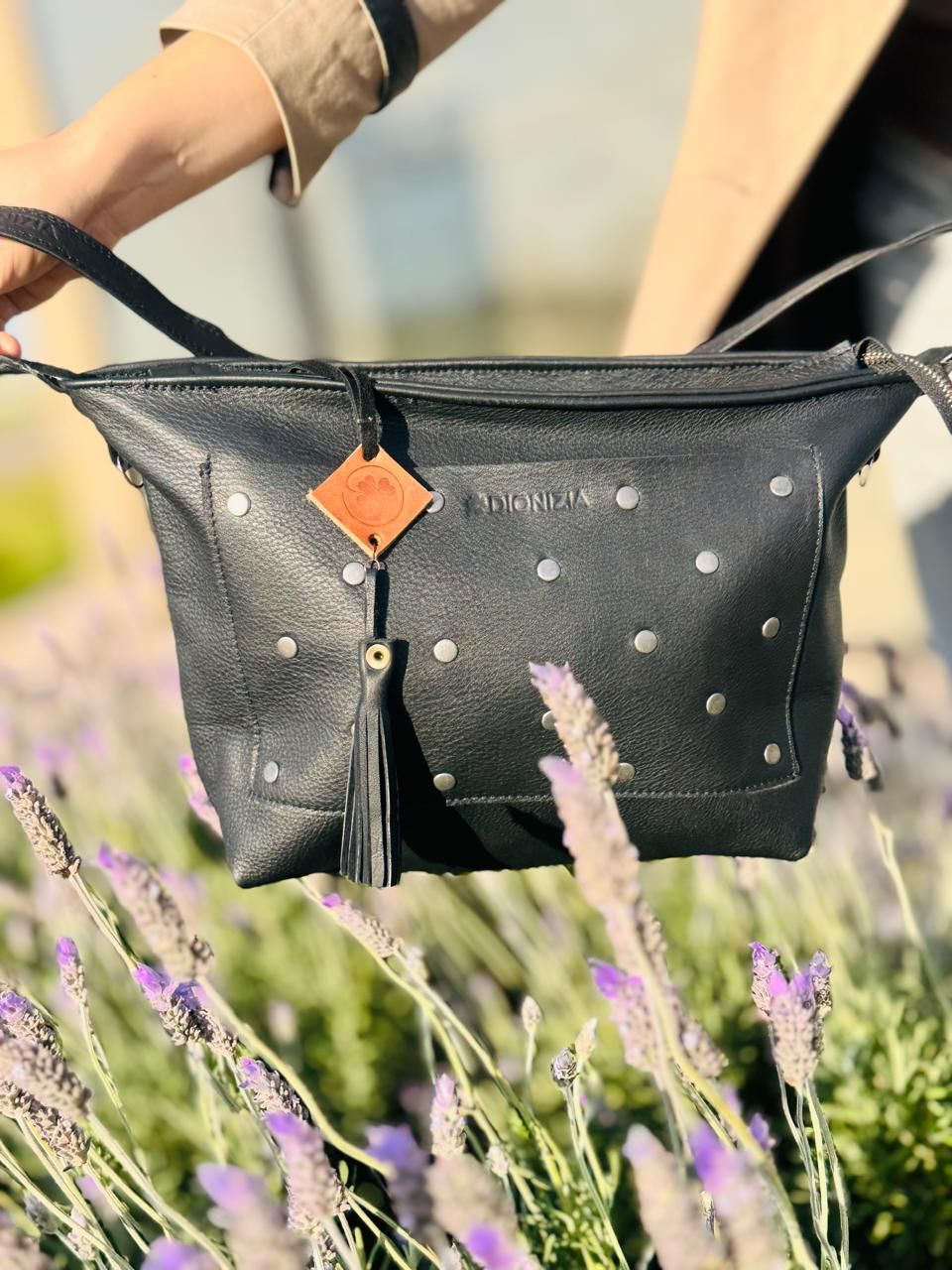 Bolso de cuero negro con tachuelas sostenido frente a flores de lavanda, con una pequeña etiqueta de cuero y borla.