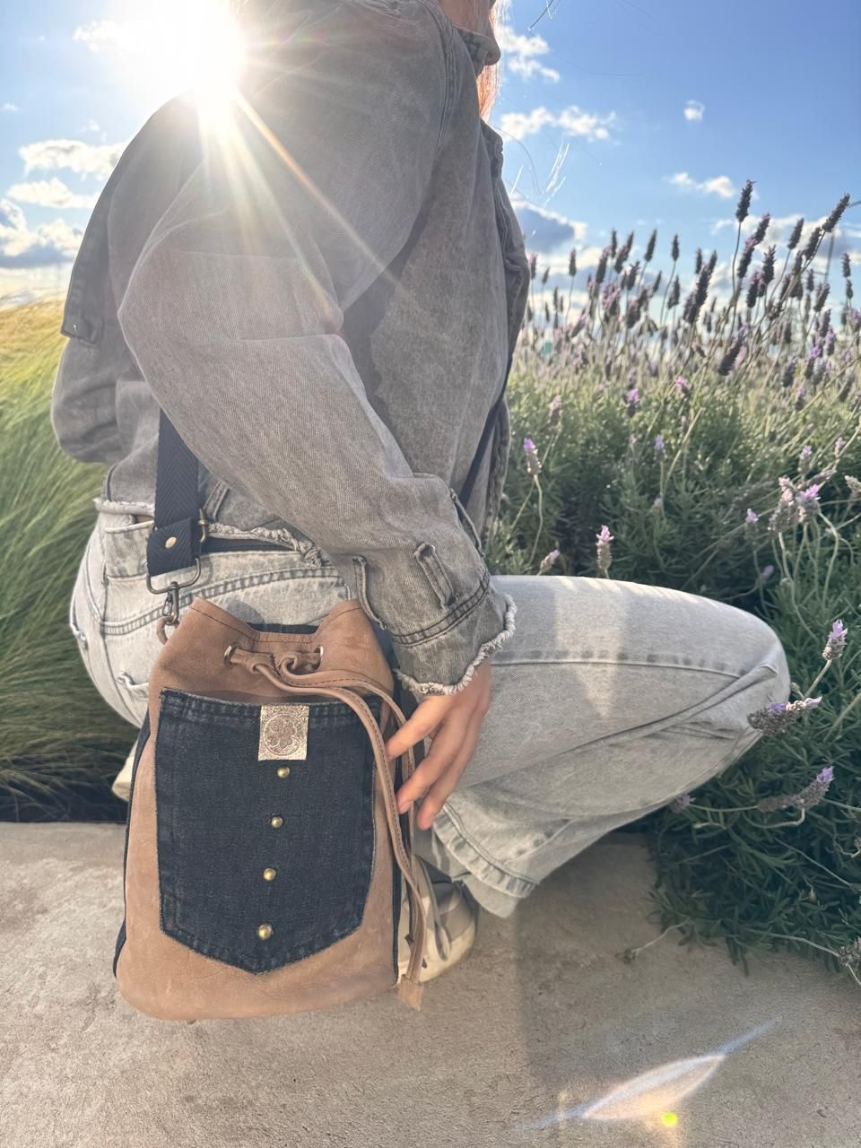 Mujer con chaqueta vaquera, agachada al aire libre, sosteniendo una bolsa de tela en un campo de lavanda. Resplandor solar.