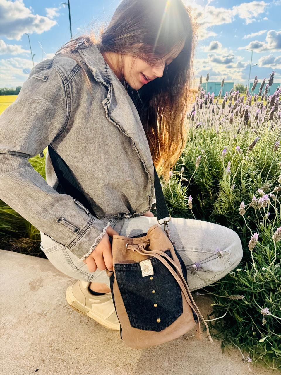 Mujer con chaqueta vaquera, agachada al aire libre, sosteniendo una bolsa de tela en un campo de lavanda. Resplandor solar.
