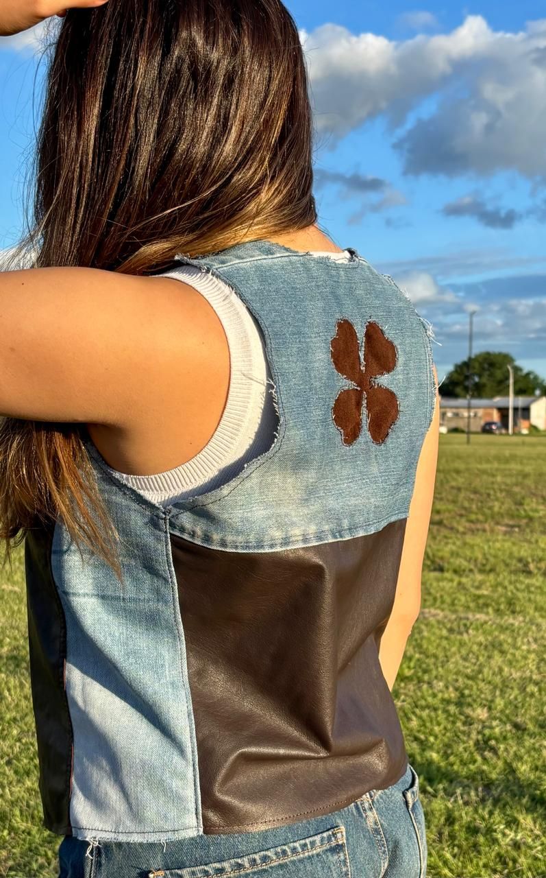 Mujer con chaleco de mezclilla y cuero en un parque, con los brazos en alto, de fondo un cielo azul.