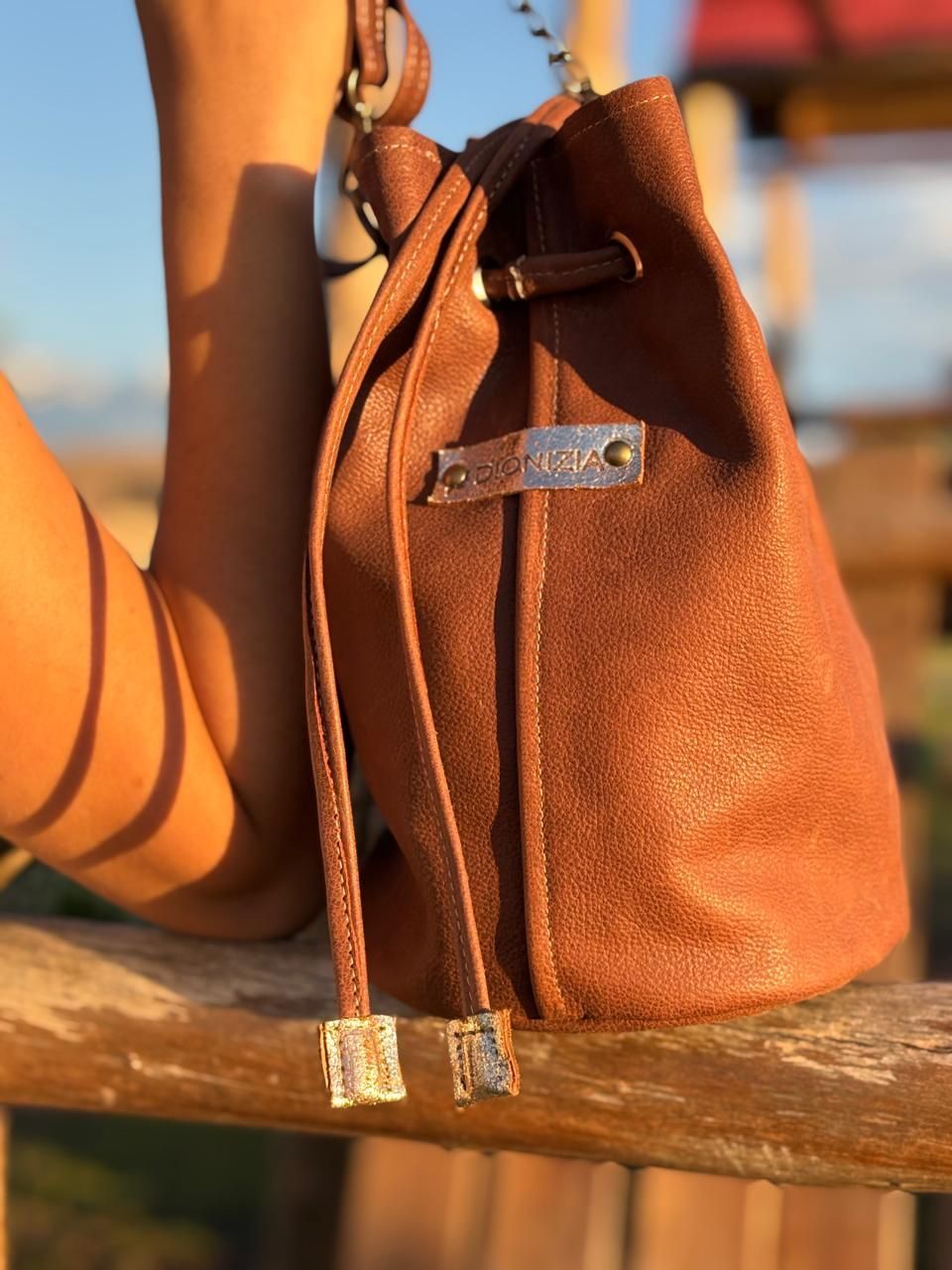 Bolsa tipo cubo de cuero marrón con cordón ajustable, apoyada sobre una barandilla de madera.