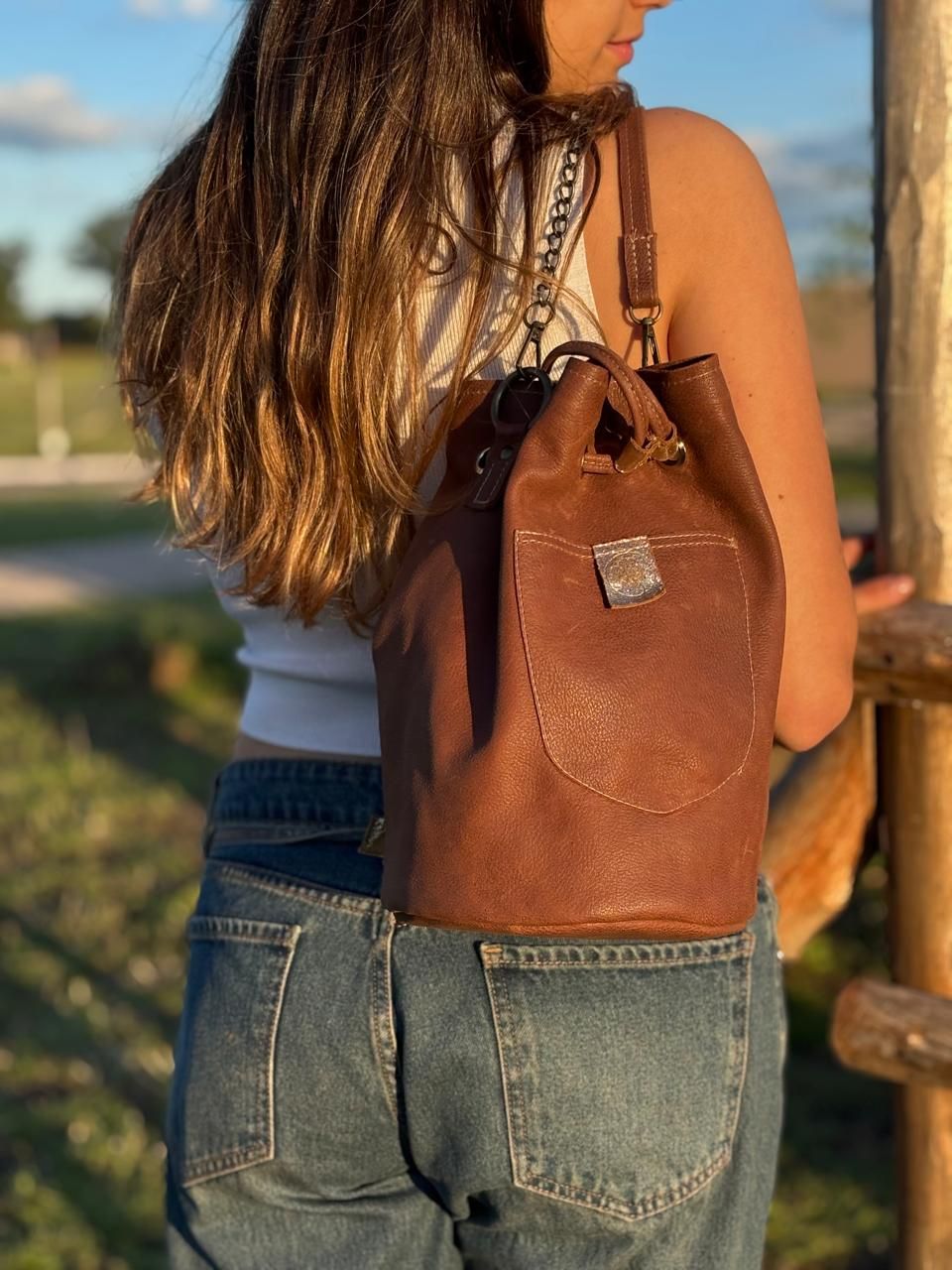 Bolsa tipo cubo de cuero marrón con cordón ajustable, apoyada sobre una barandilla de madera.