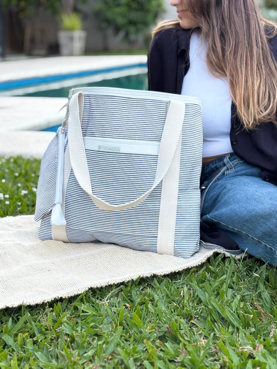 Mujer sentada en el césped con un bolso de rayas, cerca de una piscina.