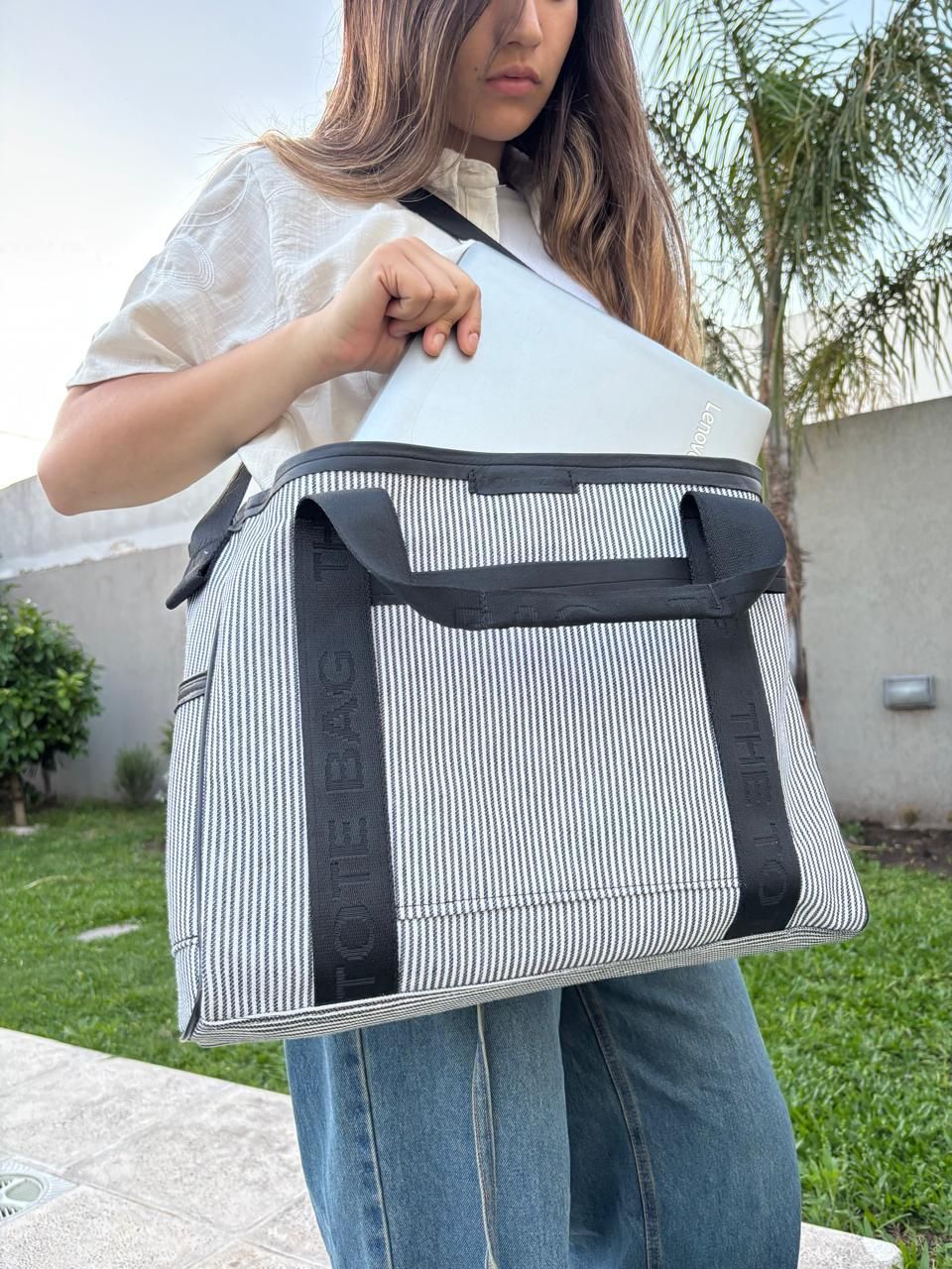 Una persona vestida con una blusa blanca y pantalones vaqueros azules sostiene un bolso tote a rayas.