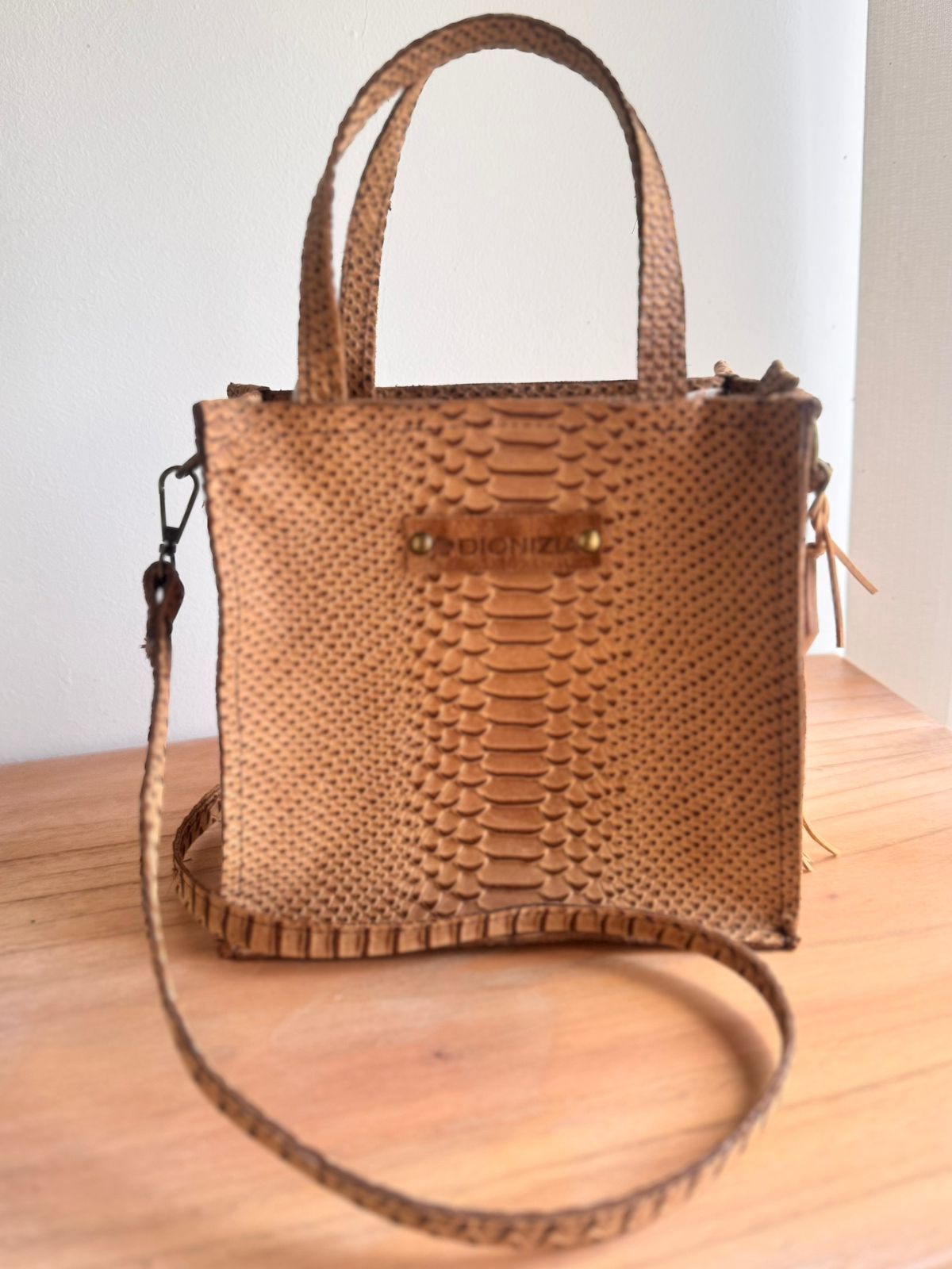 Bolso tote de piel de serpiente color canela con asas superiores y correa para el hombro sobre una superficie de madera.