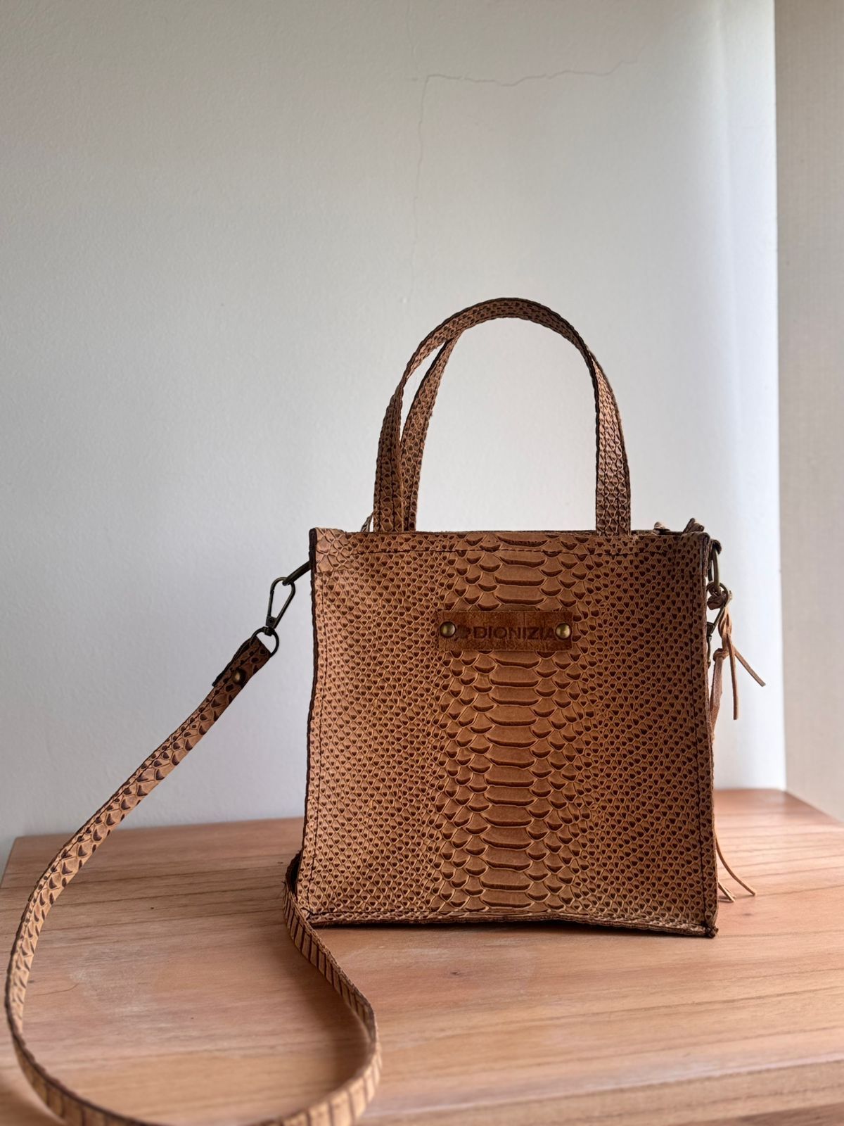 Bolso tote de piel de serpiente color canela con asas superiores y correa para el hombro sobre una superficie de madera.