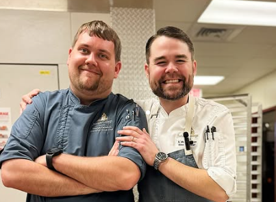 Two smiling chefs pose, arms around each other, in a kitchen setting.